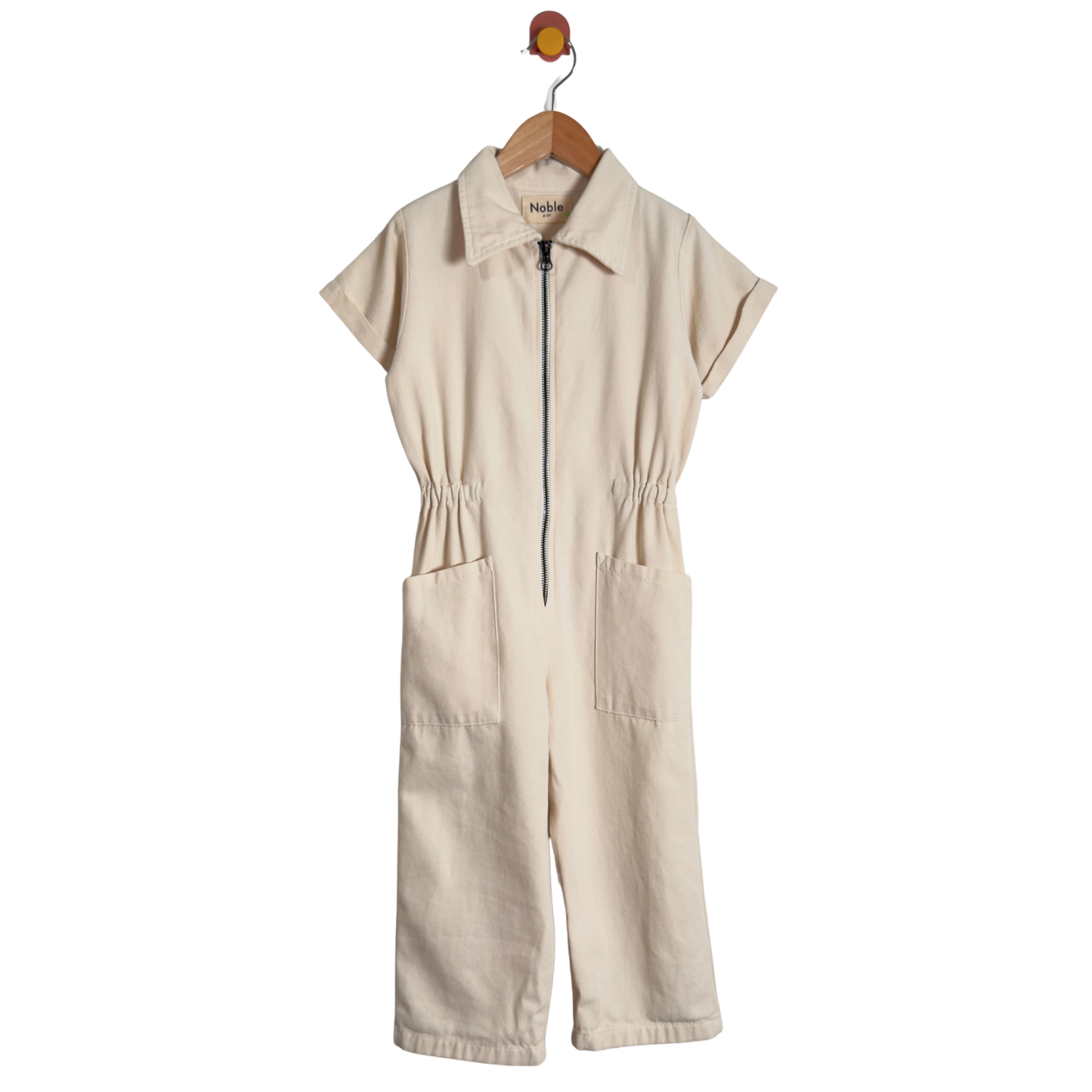 A cream-colored short-sleeve jumpsuit with a half-zip front, collar, and front patch pockets, hanging on a wooden hanger against a white background.