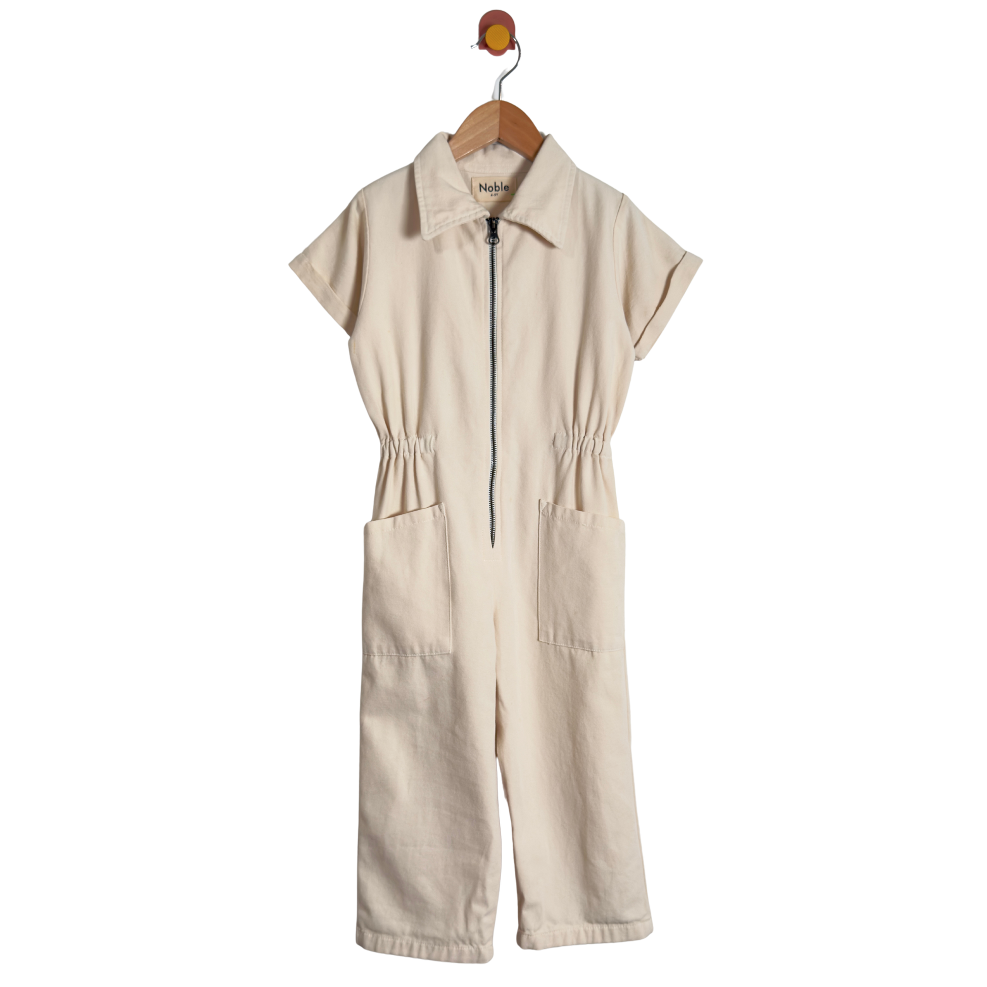 A cream-colored short-sleeve jumpsuit with a half-zip front, collar, and front patch pockets, hanging on a wooden hanger against a white background.