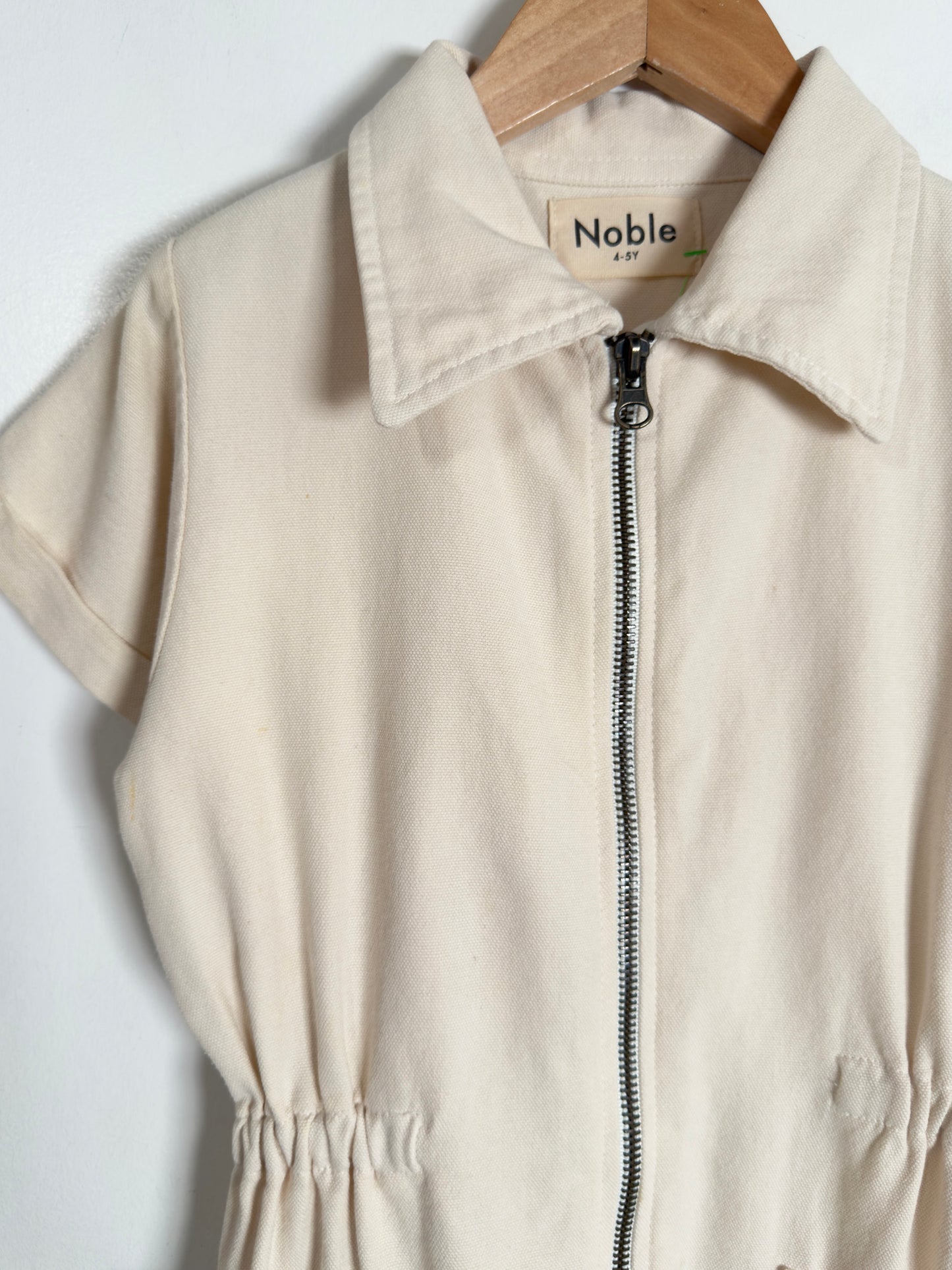 A cream-colored short-sleeve zip-up jumpsuit with a pointed collar, hanging on a wooden hanger against a plain white background. The brand label 'Noble' is visible inside the collar.