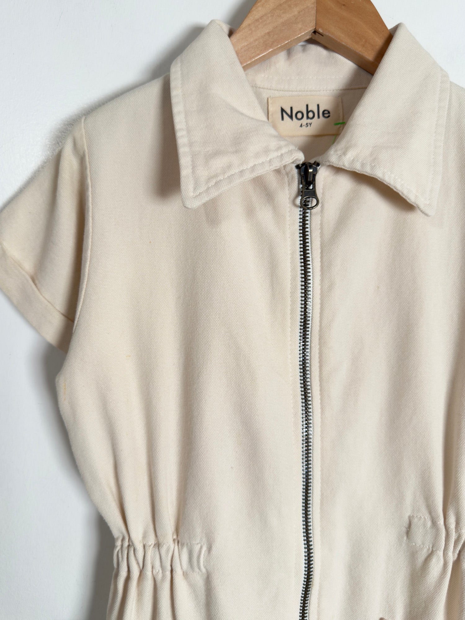 A cream-colored short-sleeve zip-up jumpsuit with a pointed collar, hanging on a wooden hanger against a plain white background. The brand label 'Noble' is visible inside the collar.