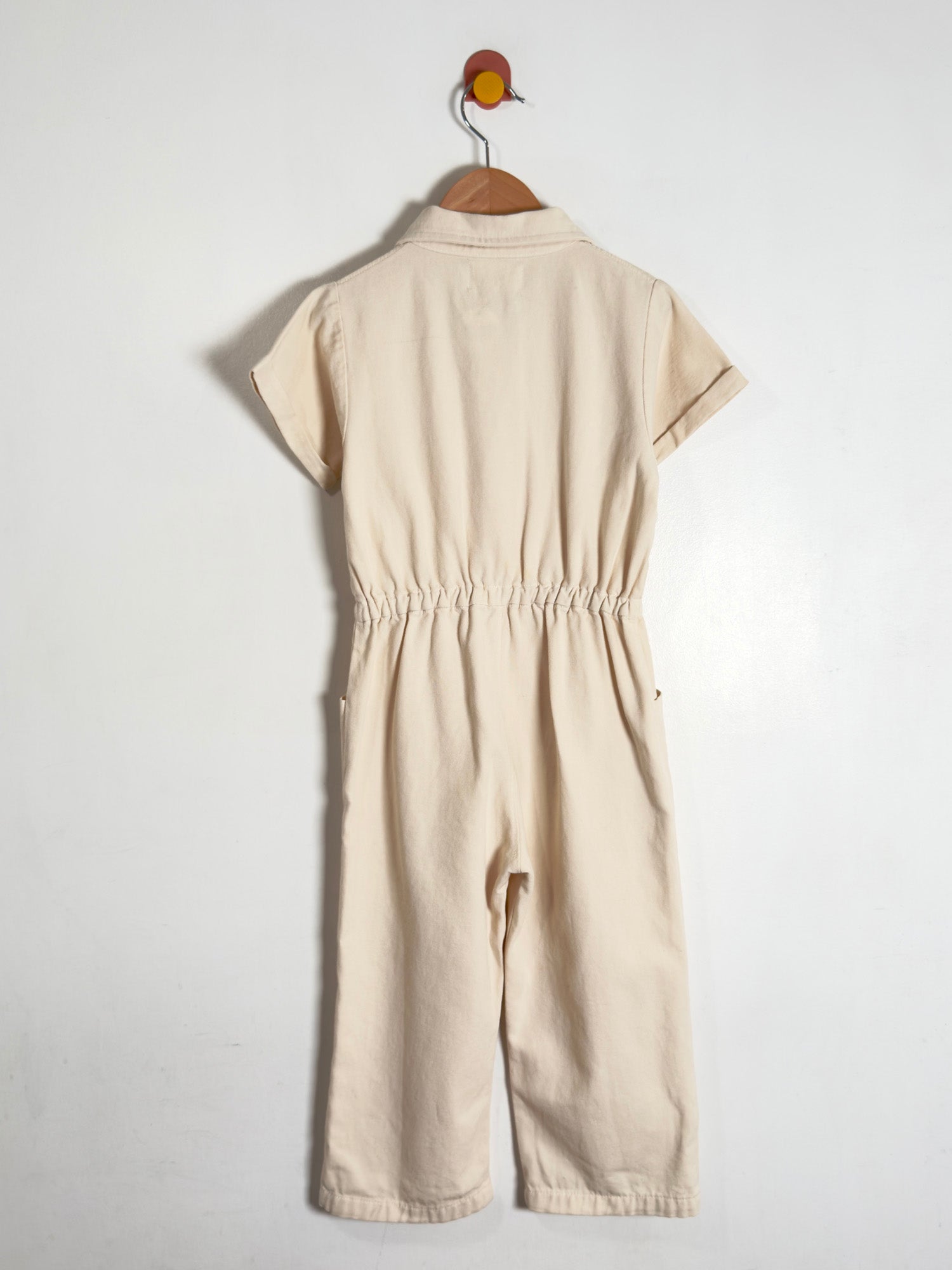 A light beige short-sleeve jumpsuit with an elastic waist, hanging on a wooden hanger against a plain white wall.