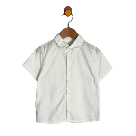 A short-sleeve button-up shirt in off-white linen fabric, hanging on a wooden hanger against a plain white background.
