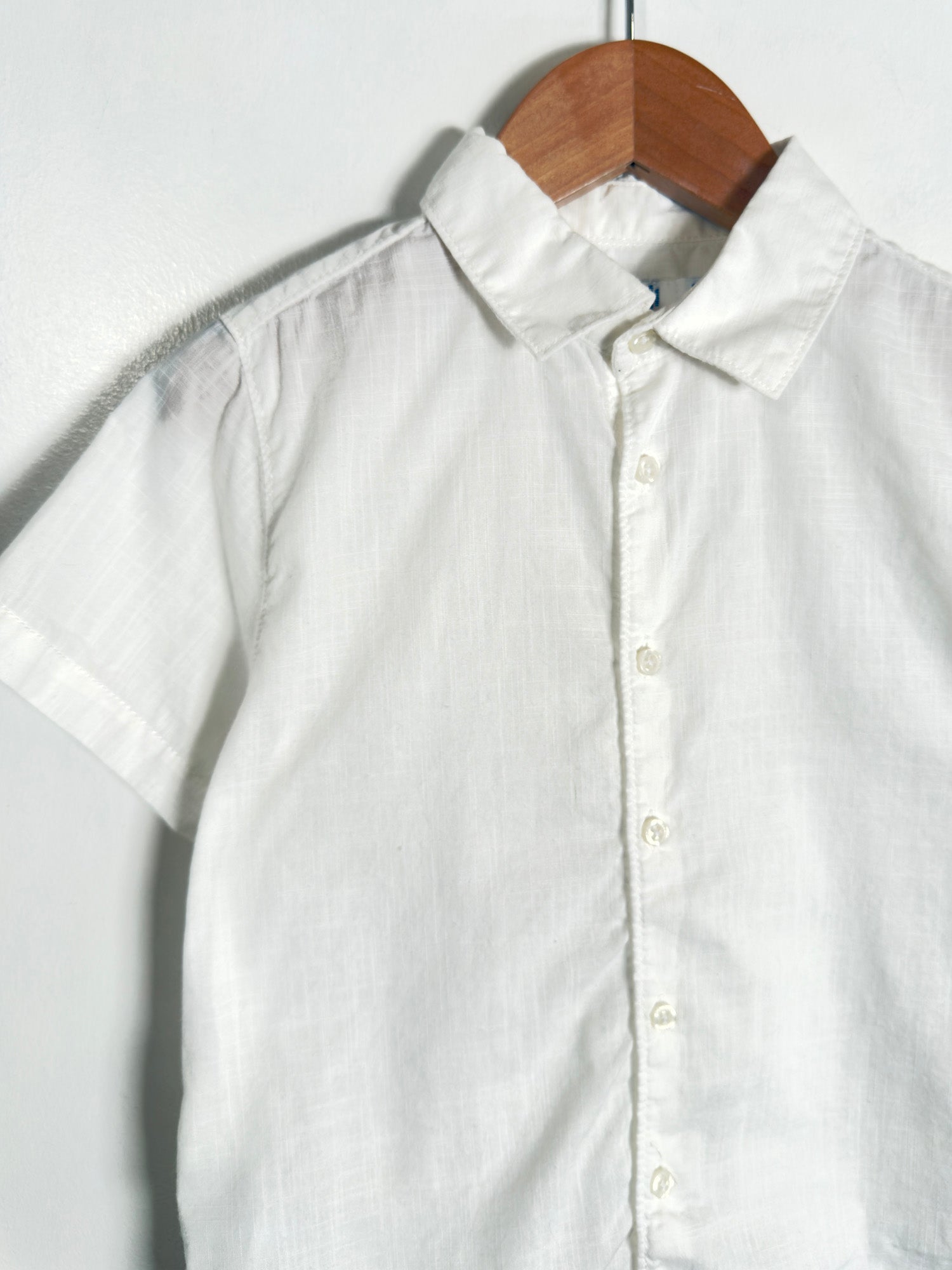 A white short-sleeve button-up shirt with a pointed collar, hanging on a wooden hanger against a plain light background.