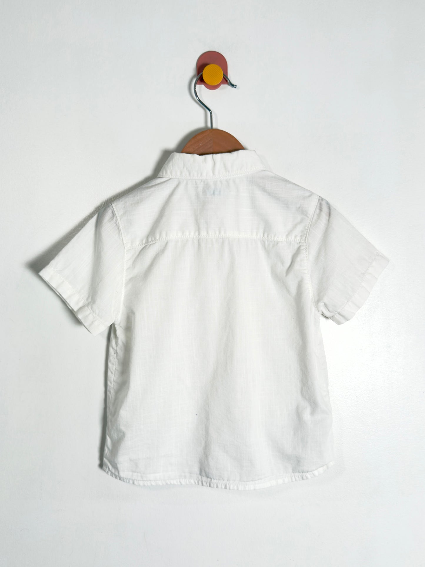 A white short-sleeve button-up shirt hanging on a wooden hanger against a plain white wall.