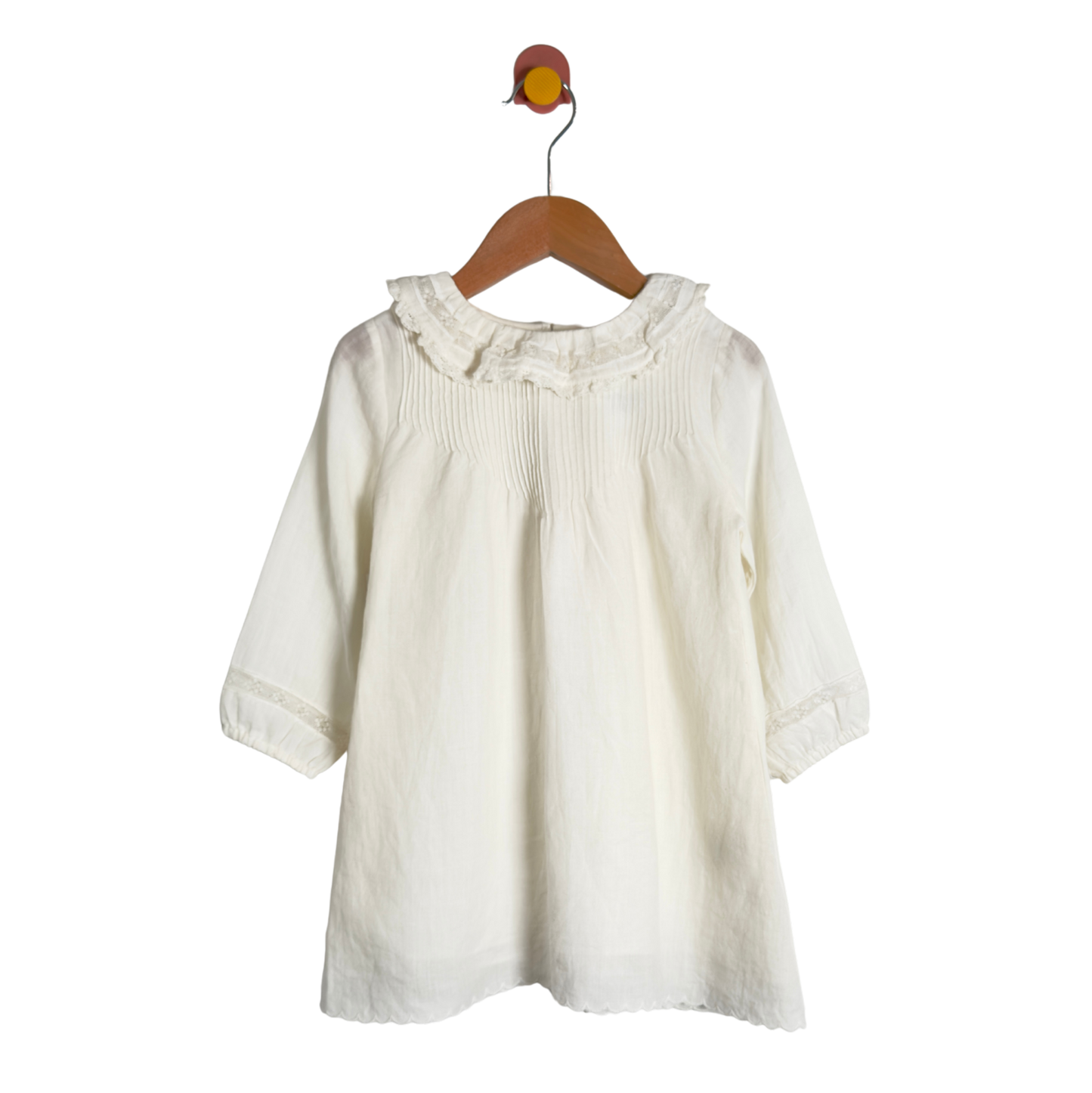 An off-white cotton blouse for toddlers, featuring a ruffled collar and smocked chest detailing, hanging on a wooden hanger against a white background.