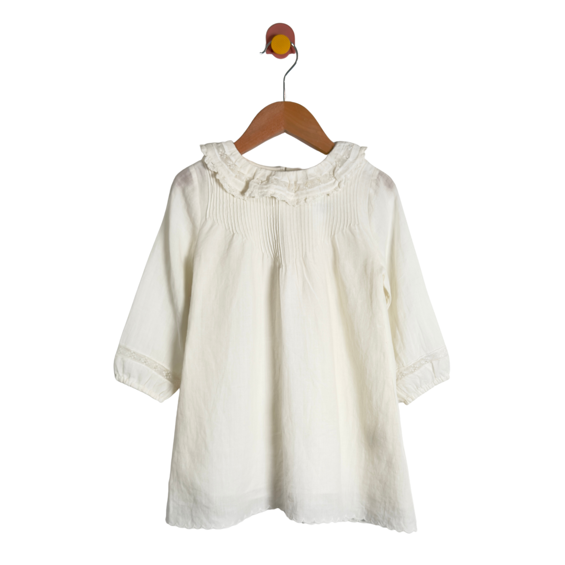 An off-white cotton blouse for toddlers, featuring a ruffled collar and smocked chest detailing, hanging on a wooden hanger against a white background.