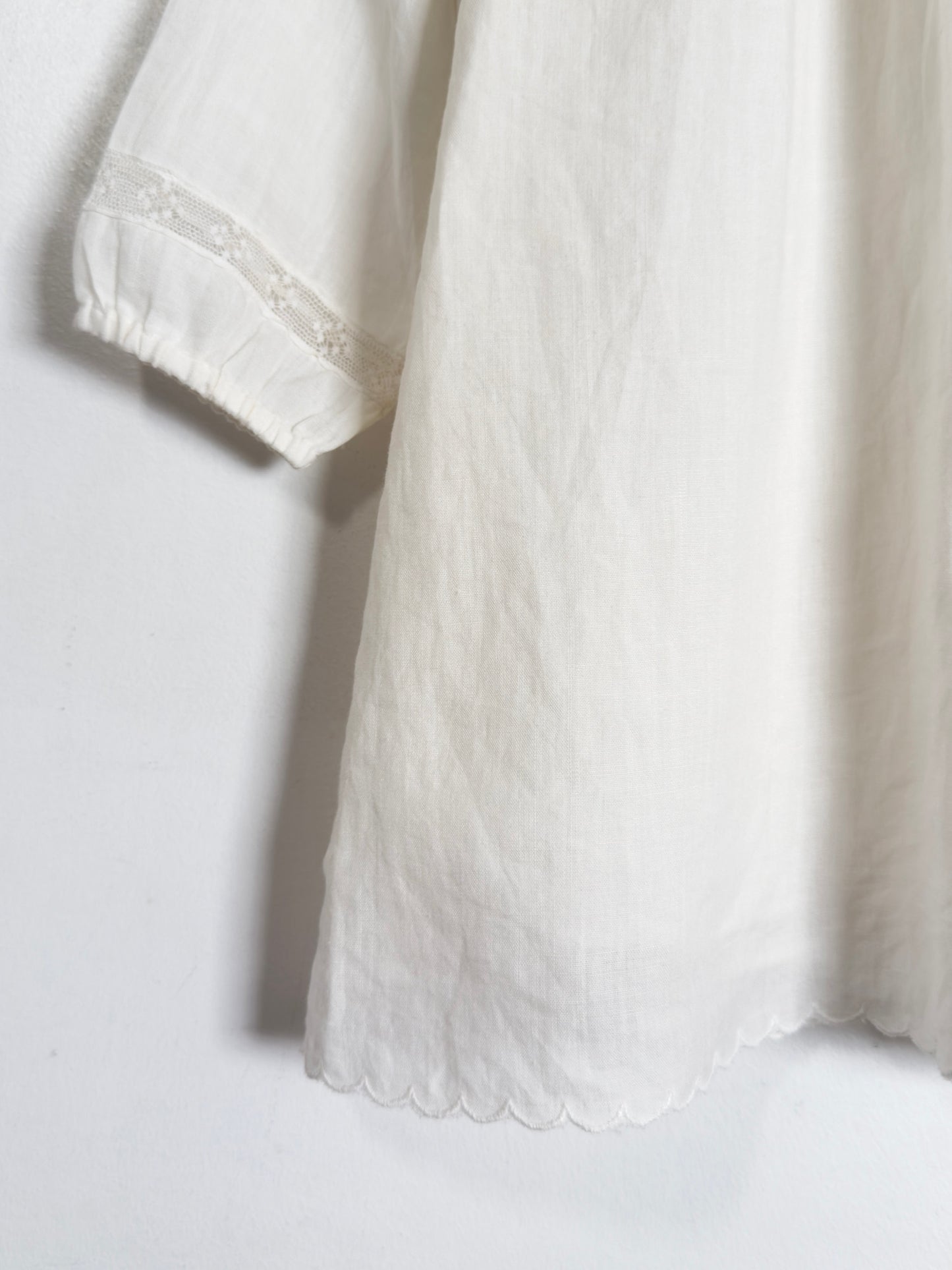 Close-up of a white cotton or linen garment with a scalloped hem and a lace-trimmed cuff, shown against a plain white background.