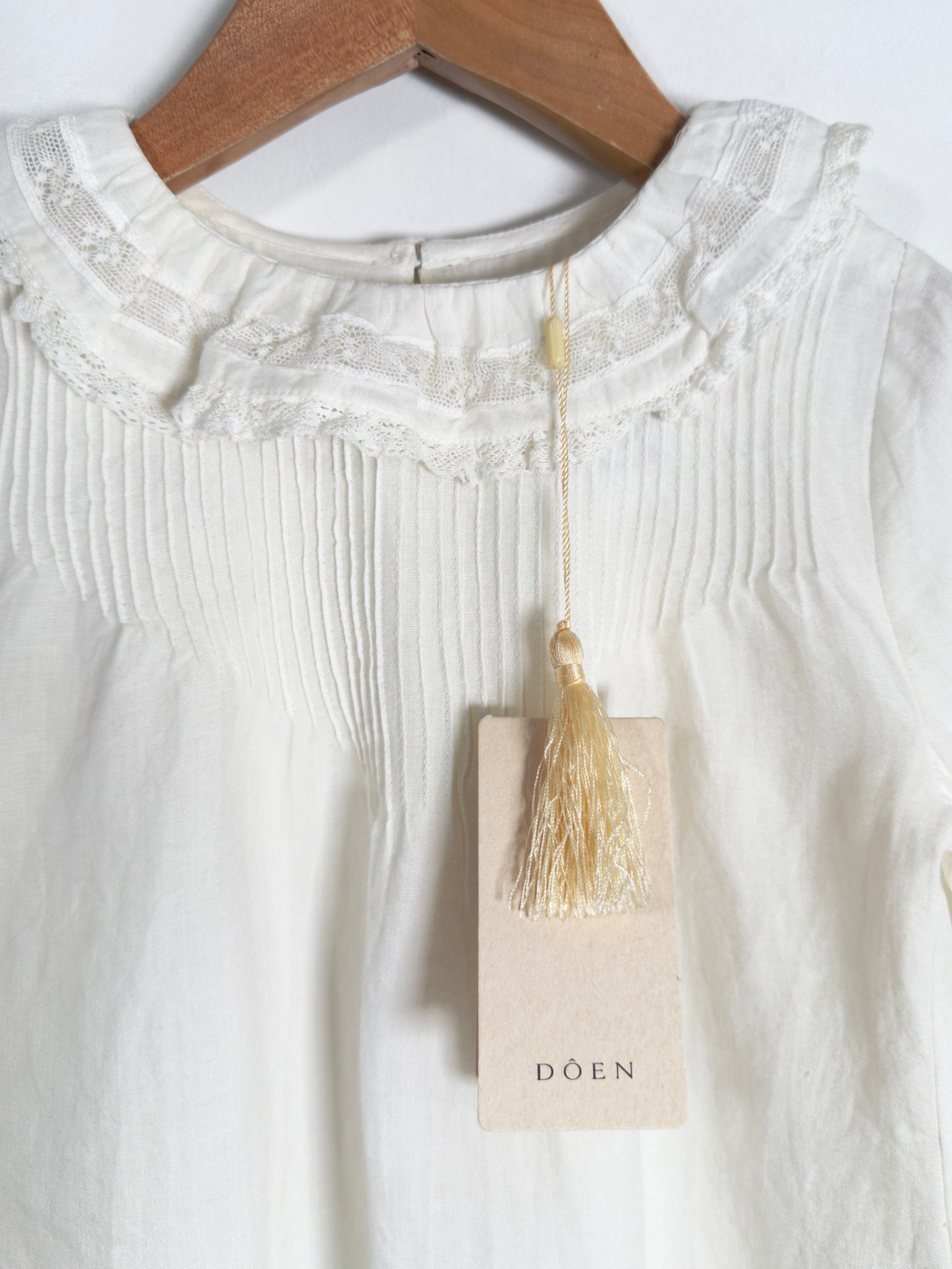 White long-sleeve blouse with a pleated bodice and lace ruffled collar, hanging on a wooden hanger with a beige brand tag that reads DOEN.