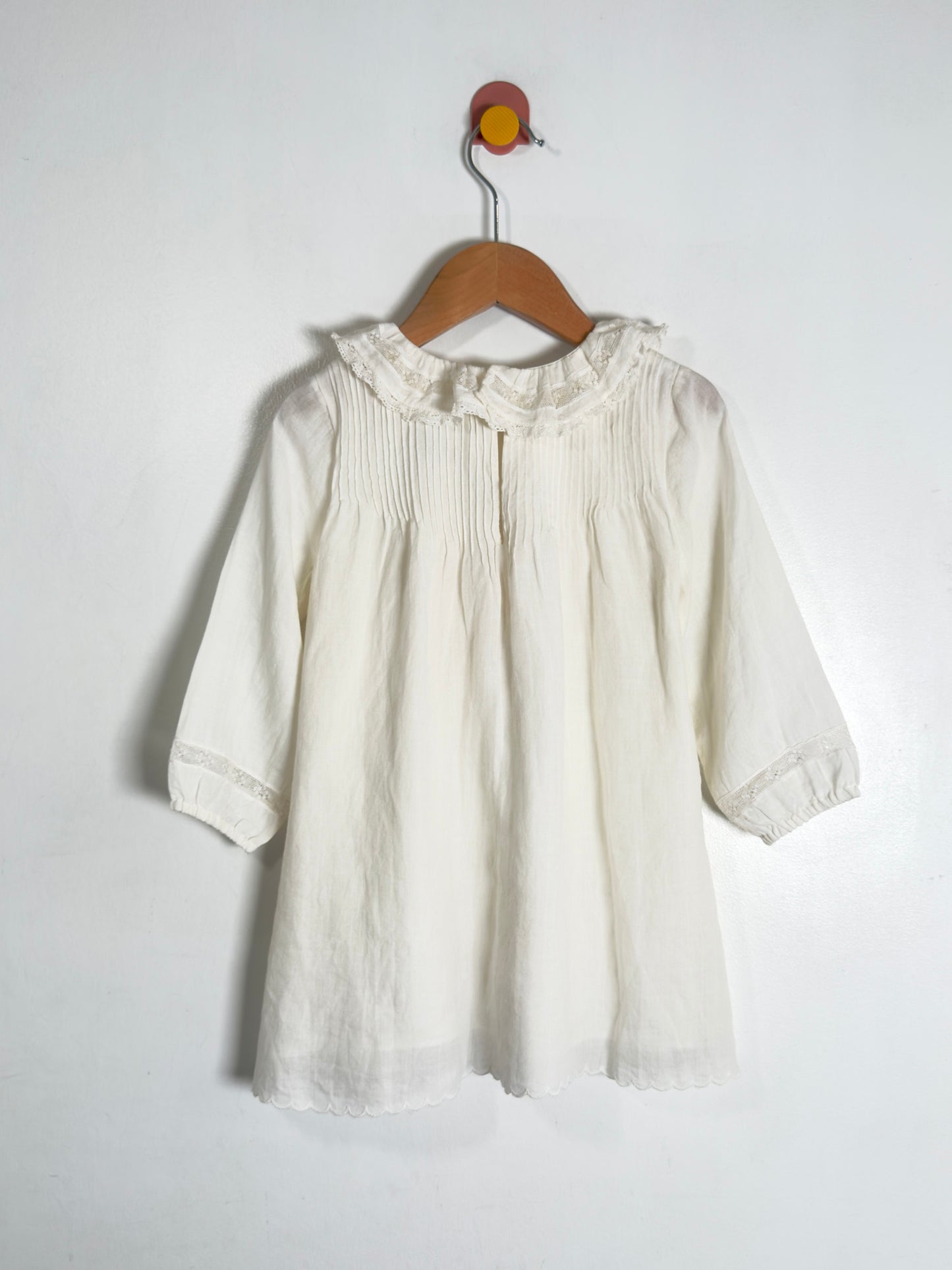 An off-white cotton blouse with a ruffled collar and smocked chest detailing, hanging on a wooden hanger against a plain white wall.