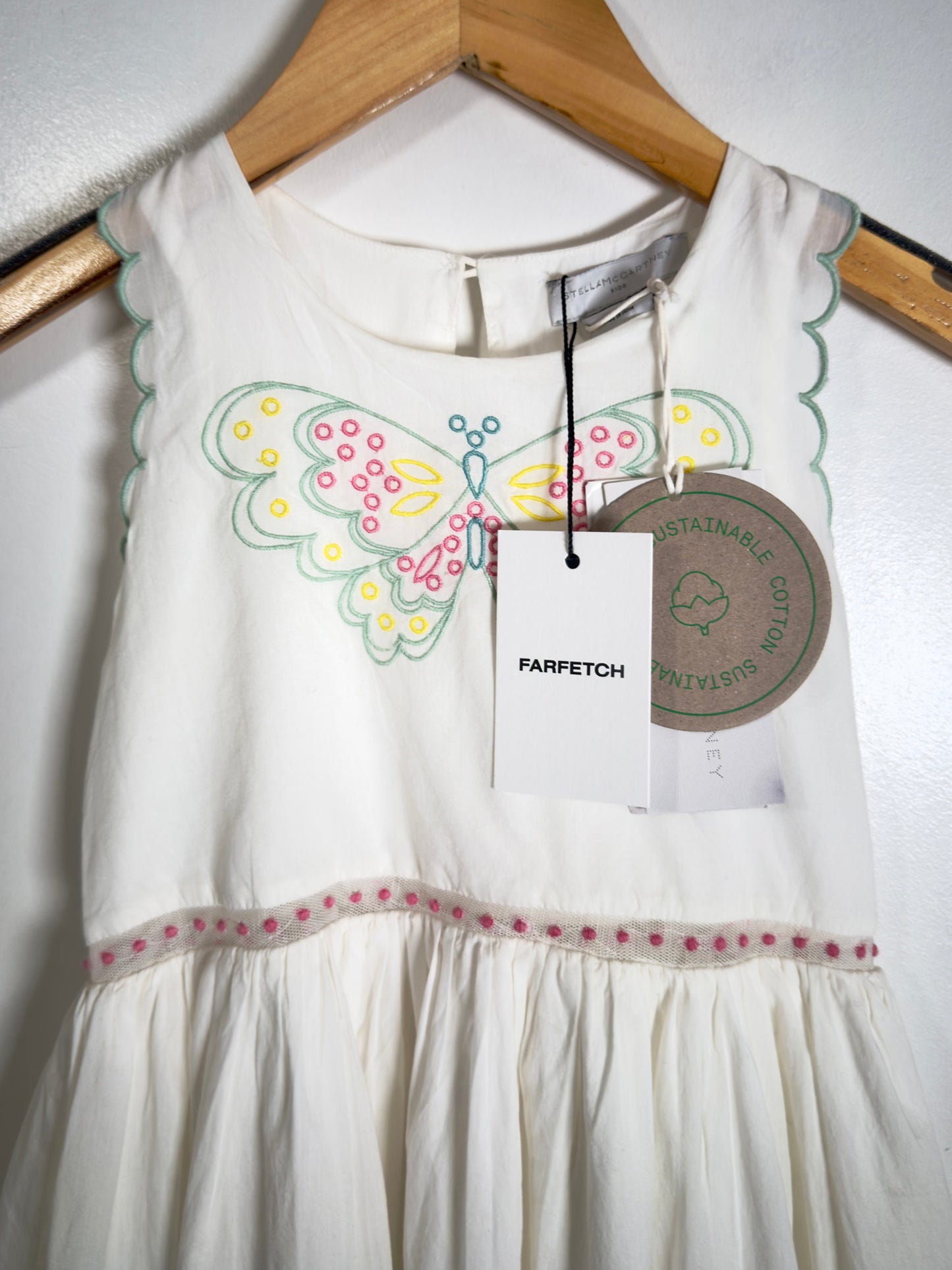 A white sleeveless baby dress with scalloped edges, featuring a colorful embroidered butterfly on the chest and a pink polka dot trim at the waist. The dress is hanging on a wooden hanger with a Parfetch brand tag and a circular sustainability tag against a white wall.