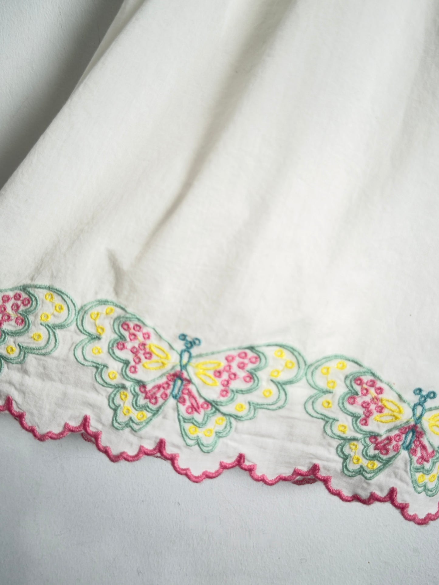 A close-up of a white fabric featuring a scalloped border with embroidered butterflies in yellow, pink, and green.