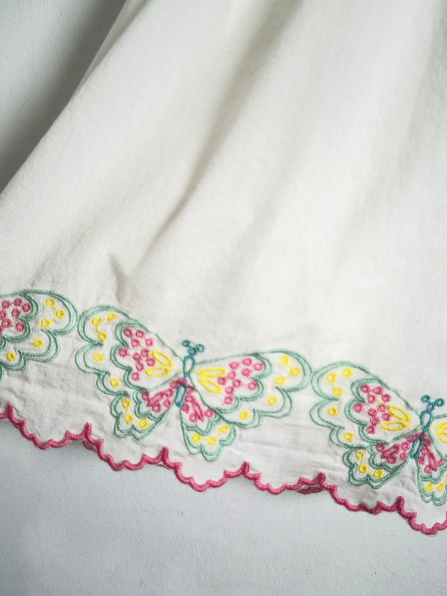 A close-up of a white fabric featuring a scalloped border with embroidered butterflies in yellow, pink, and green.