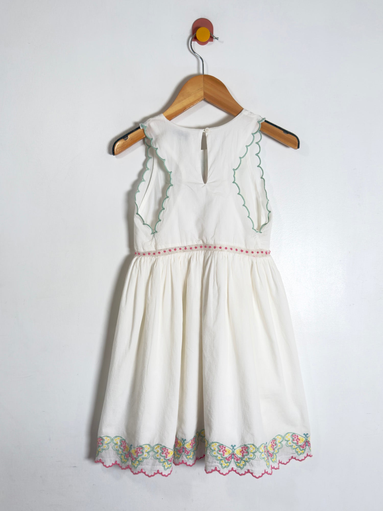 An off-white sleeveless baby dress with green embroidered details and a floral lace hem, hanging on a wooden hanger against a plain white wall.