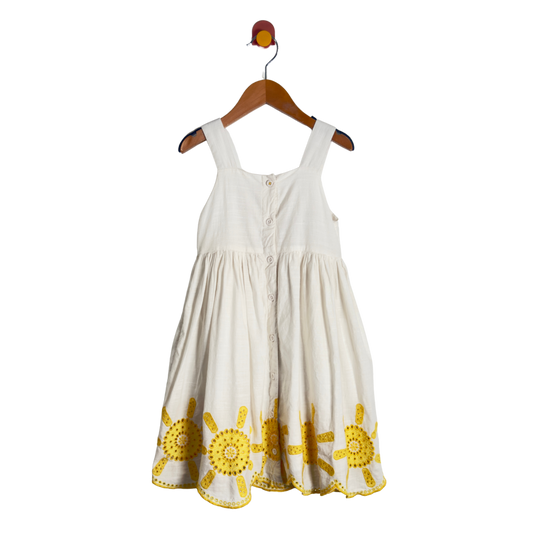 An off-white sleeveless baby dress with yellow sun embroidery along the hem, hanging on a wooden hanger against a white background.
