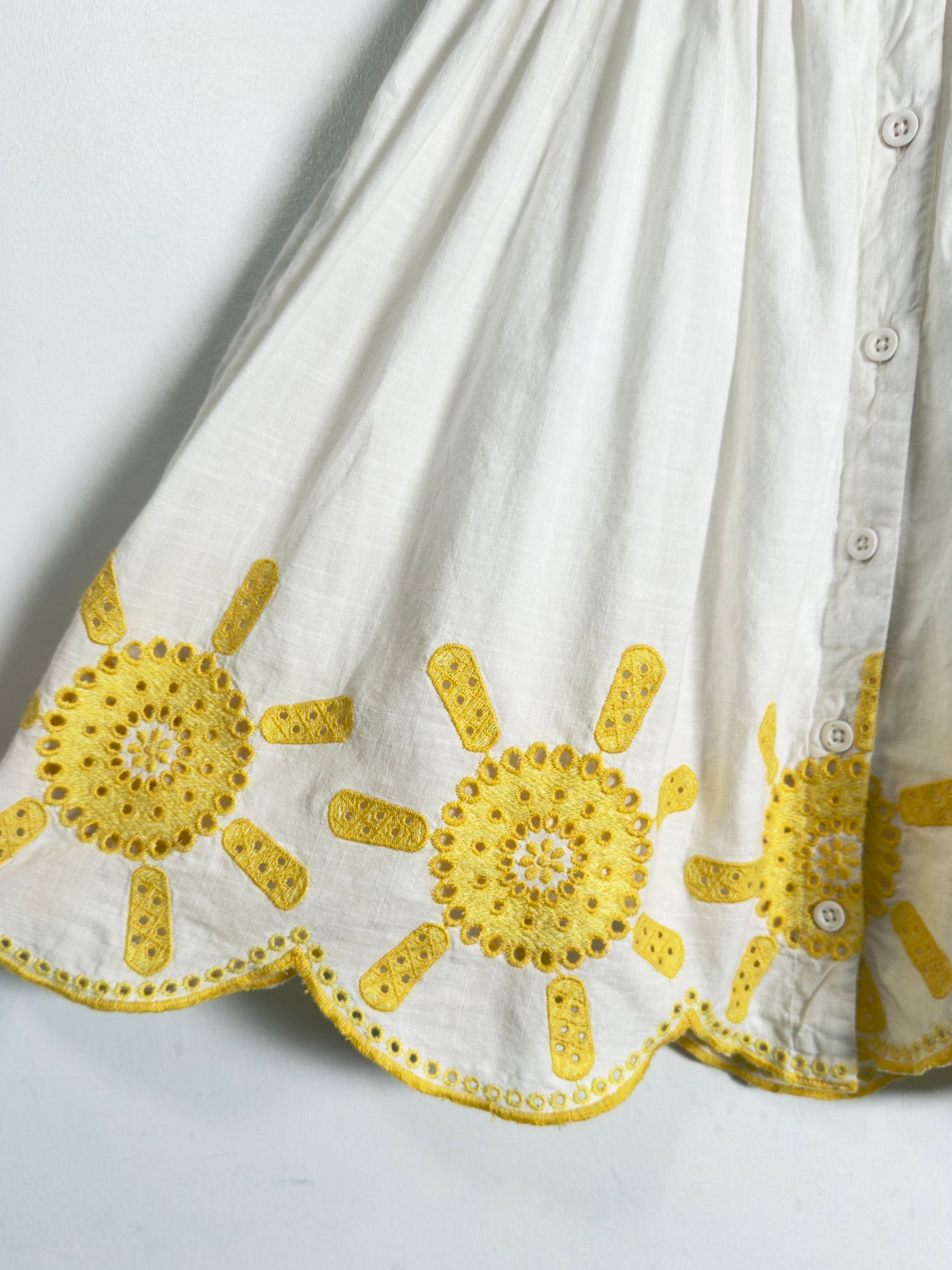 Close-up of a white fabric garment with yellow embroidered sunburst motifs and a scalloped hem, showing a buttoned front edge against a plain light background.