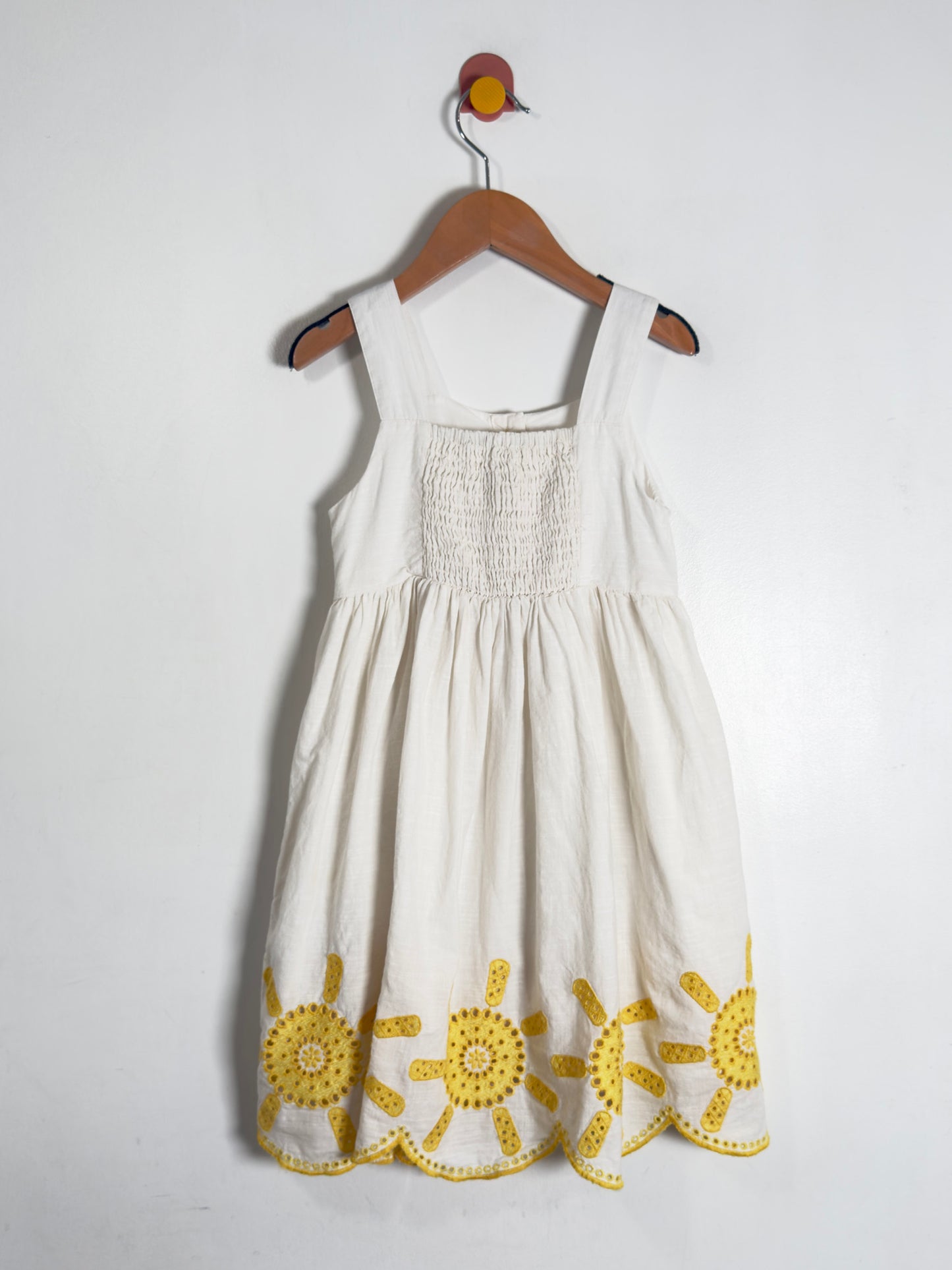 A white sleeveless baby dress with yellow embroidered sun motifs at the hem, hanging on a wooden hanger against a plain white wall.