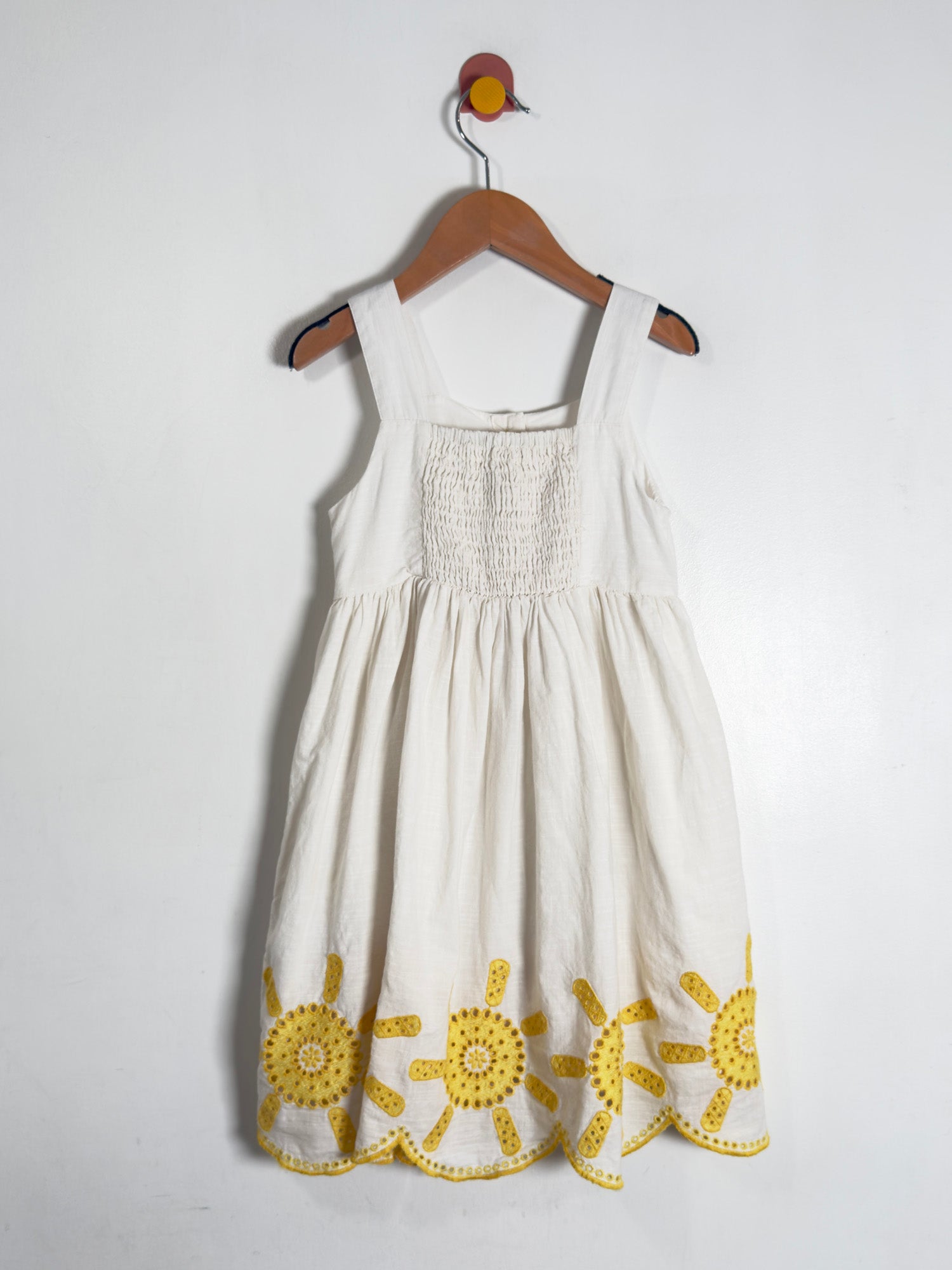 A white sleeveless baby dress with yellow embroidered sun motifs at the hem, hanging on a wooden hanger against a plain white wall.