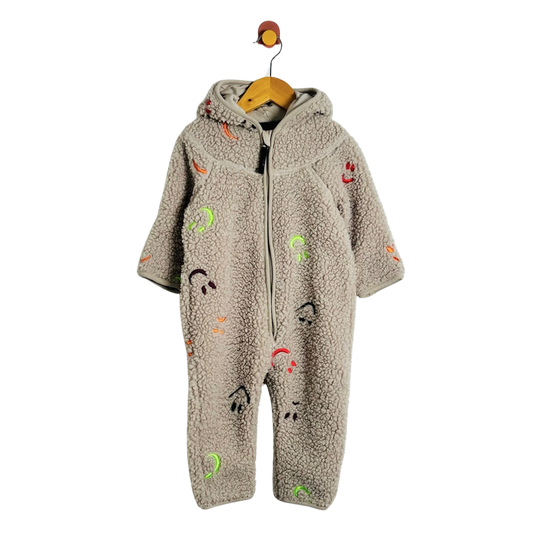 A baby hooded one-piece fleece jumpsuit in a warm taupe-beige color, decorated with colorful cartoon face prints, hanging on a wooden hanger against a plain white background.