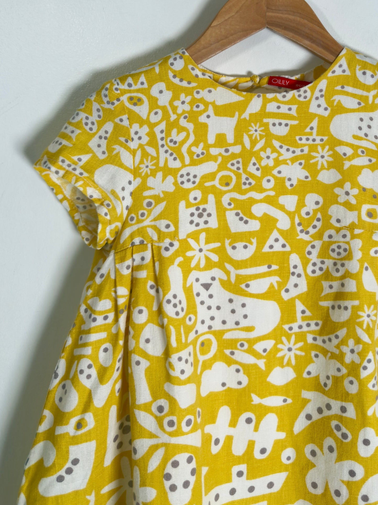 A yellow short-sleeve dress with a white abstract pattern of animals, stars, and shapes, hanging on a wooden hanger against a plain white wall.