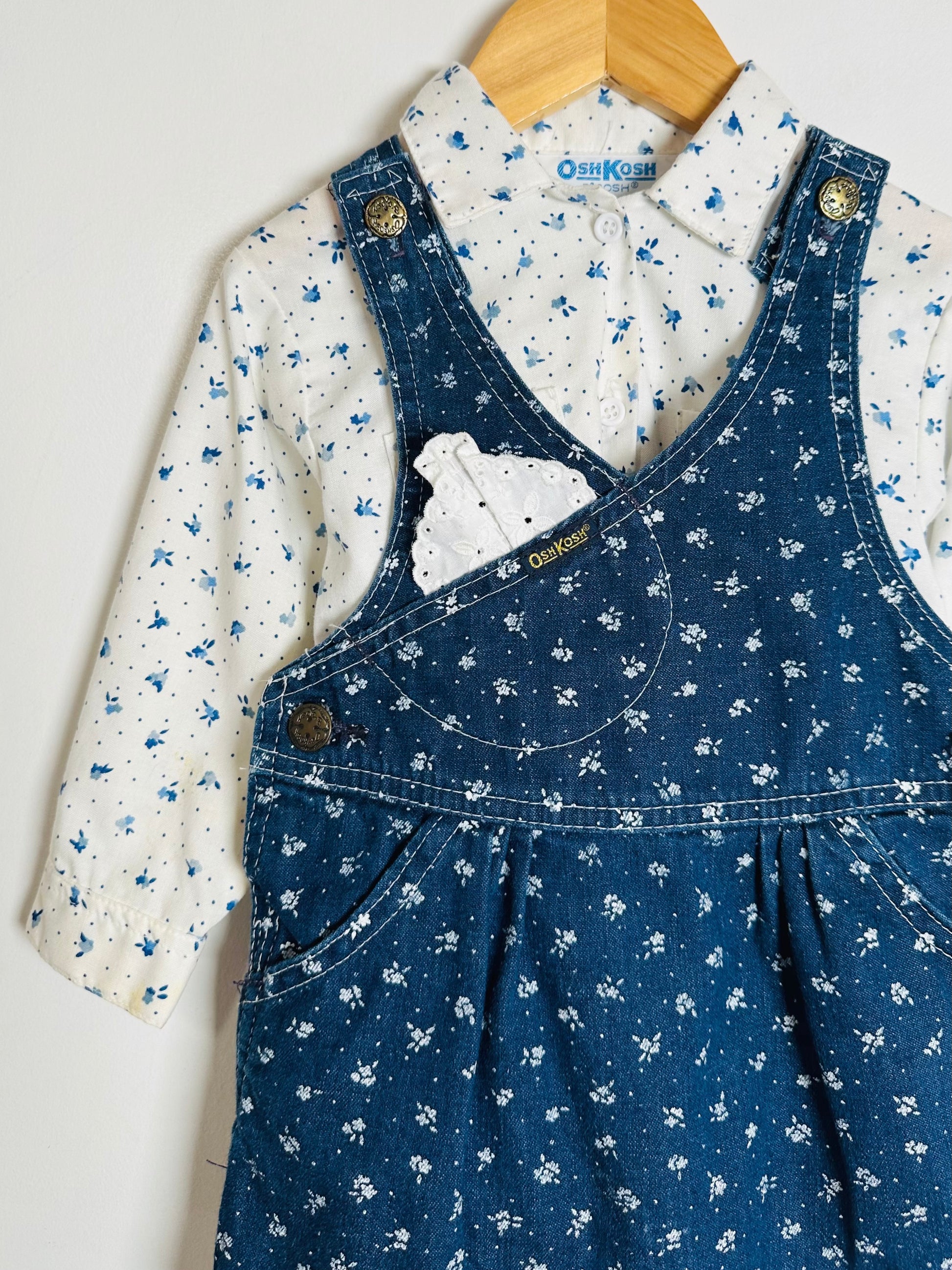 Blue floral overalls with a white shirt on a wooden hanger against a light gray background