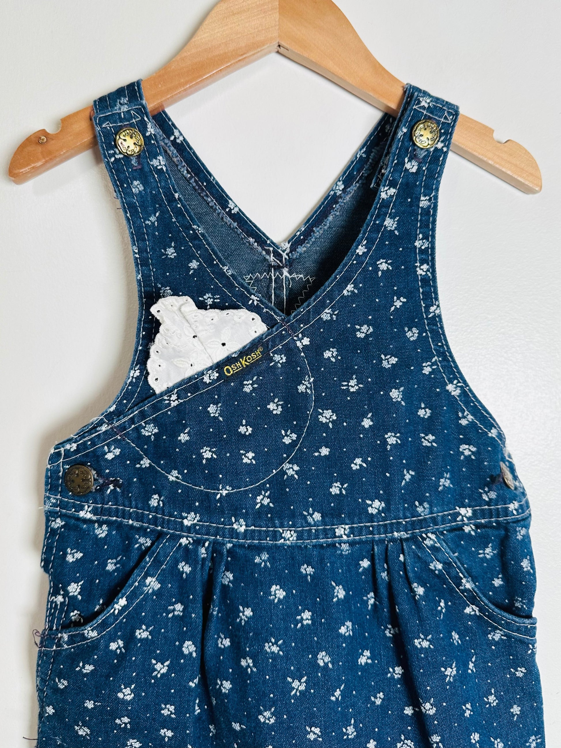 Blue denim overalls with white floral pattern on a hanger against a white background