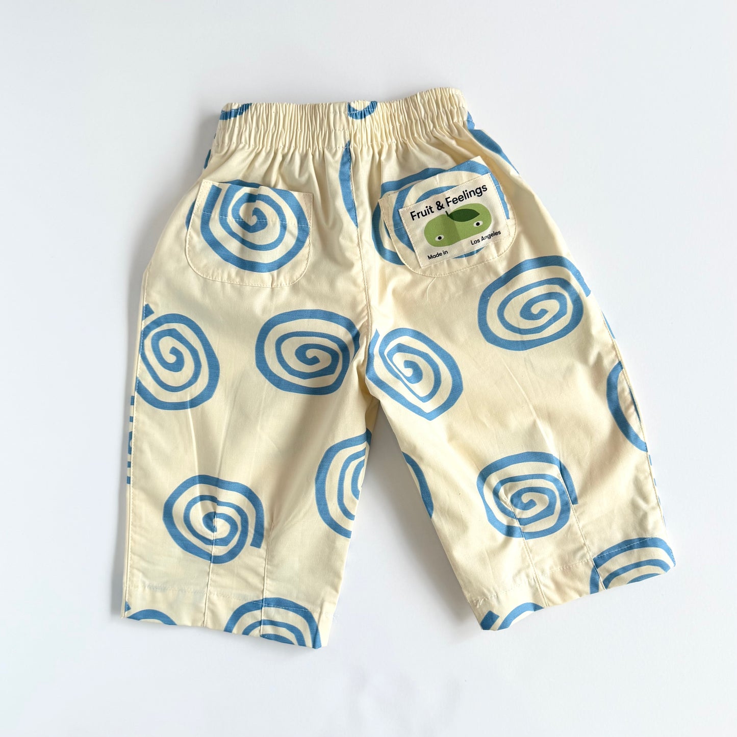A pair of cream-colored pants with a blue spiral pattern, featuring an elastic waistband and a side patch that reads 'Fruit & Feelings'.