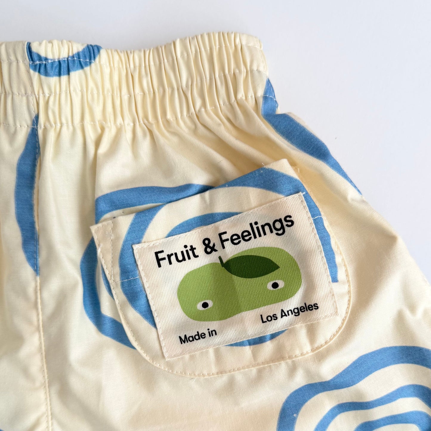 Close-up of cream-colored baby pants with a blue spiral pattern, featuring a pocket with a patch that reads 'Fruit & Feelings Made in Los Angeles' and shows a green fruit illustration.