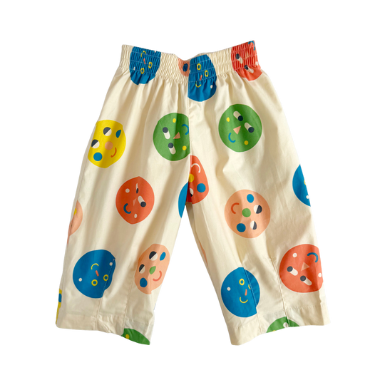 A pair of cream-colored children's pants with a colorful pattern of cartoonish smiling faces in blue, green, orange, and yellow, featuring an elastic waistband.