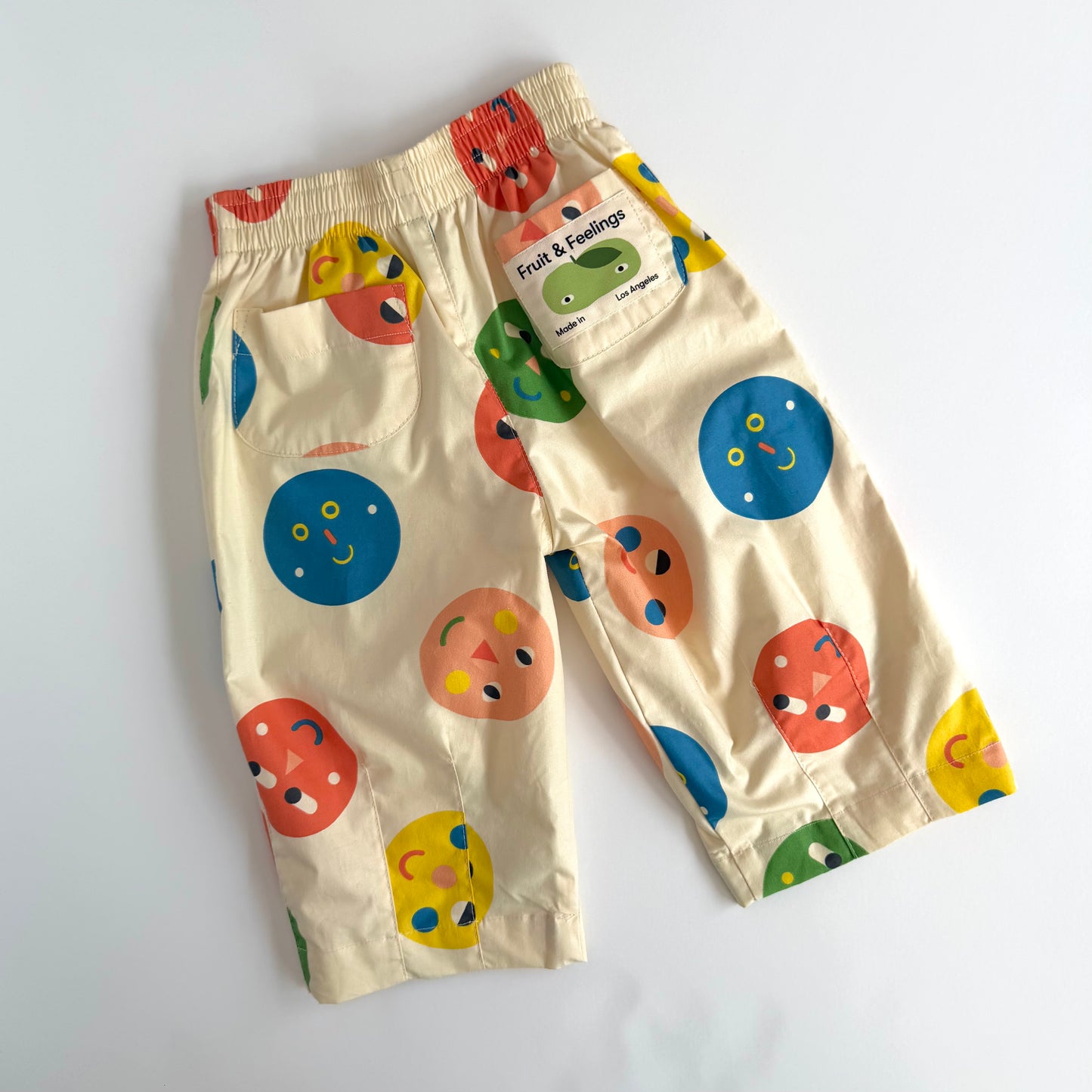 A pair of cream-colored children's pants with a colorful fruit and vegetable face print, featuring a side pocket with a 'Fruit & Feelings' brand tag, laid flat on a white background.