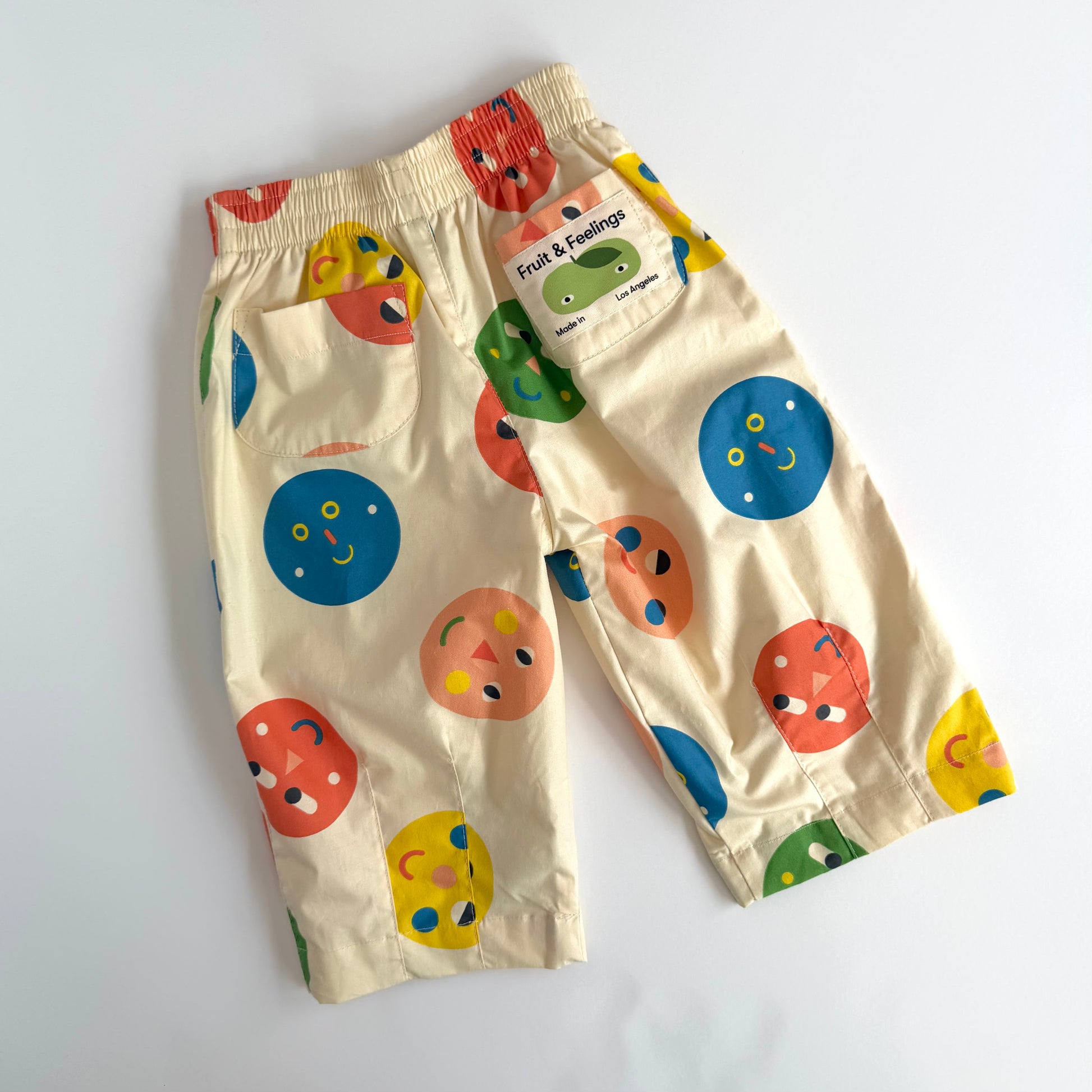 A pair of cream-colored children's pants with a colorful fruit and vegetable face print, featuring a side pocket with a 'Fruit & Feelings' brand tag, laid flat on a white background.