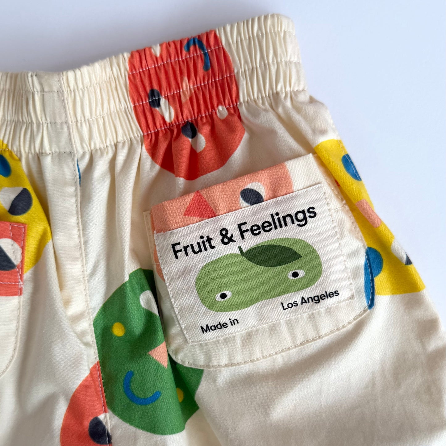 Close-up of a pair of cream-colored children's pants with a colorful fruit and abstract pattern, featuring a sewn-on patch that reads 'Fruit & Feelings' with a green leaf character and 'Made in Los Angeles'.