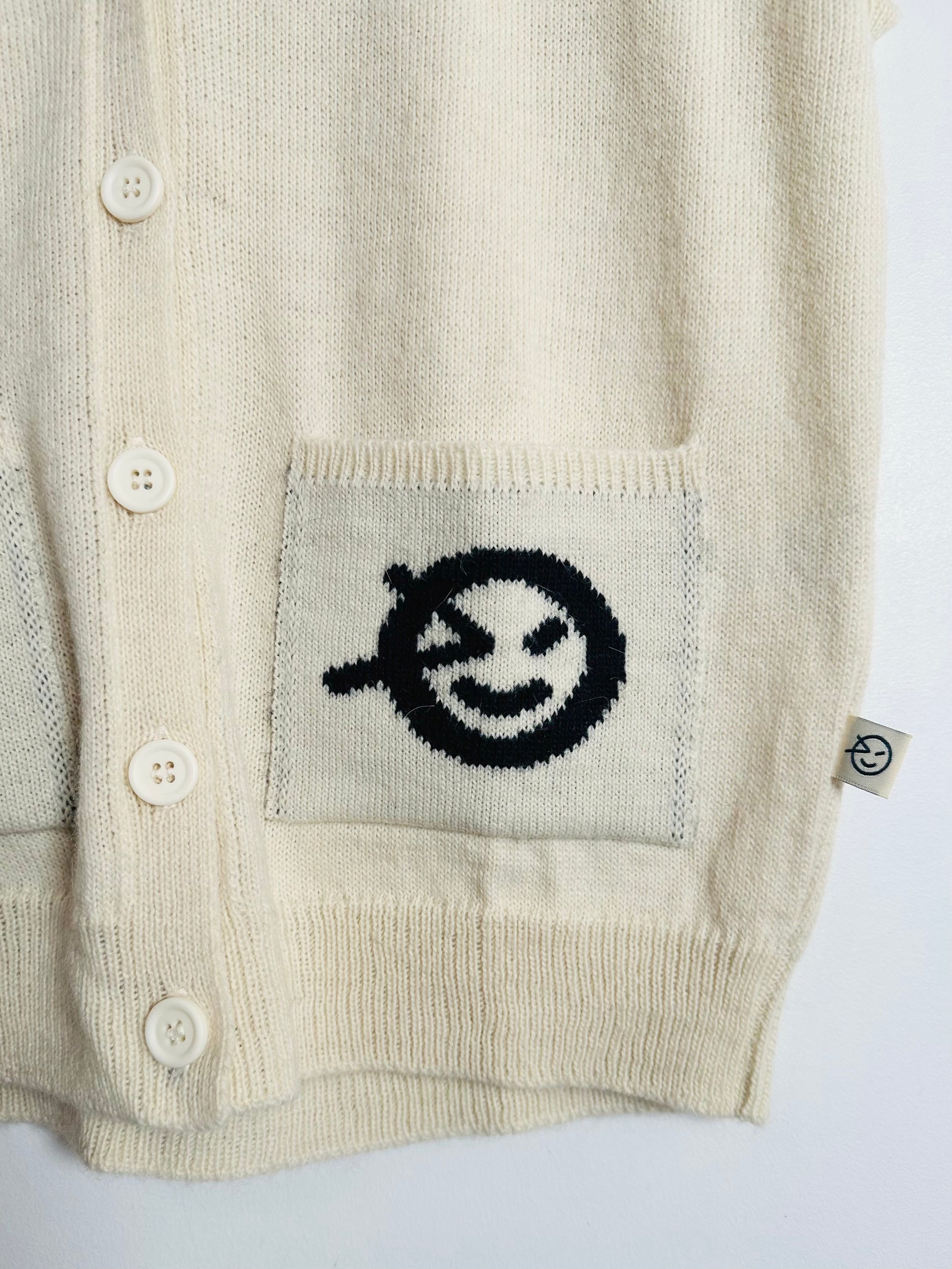 Beige cardigan with a black and white embroidered design on the pocket against a white background