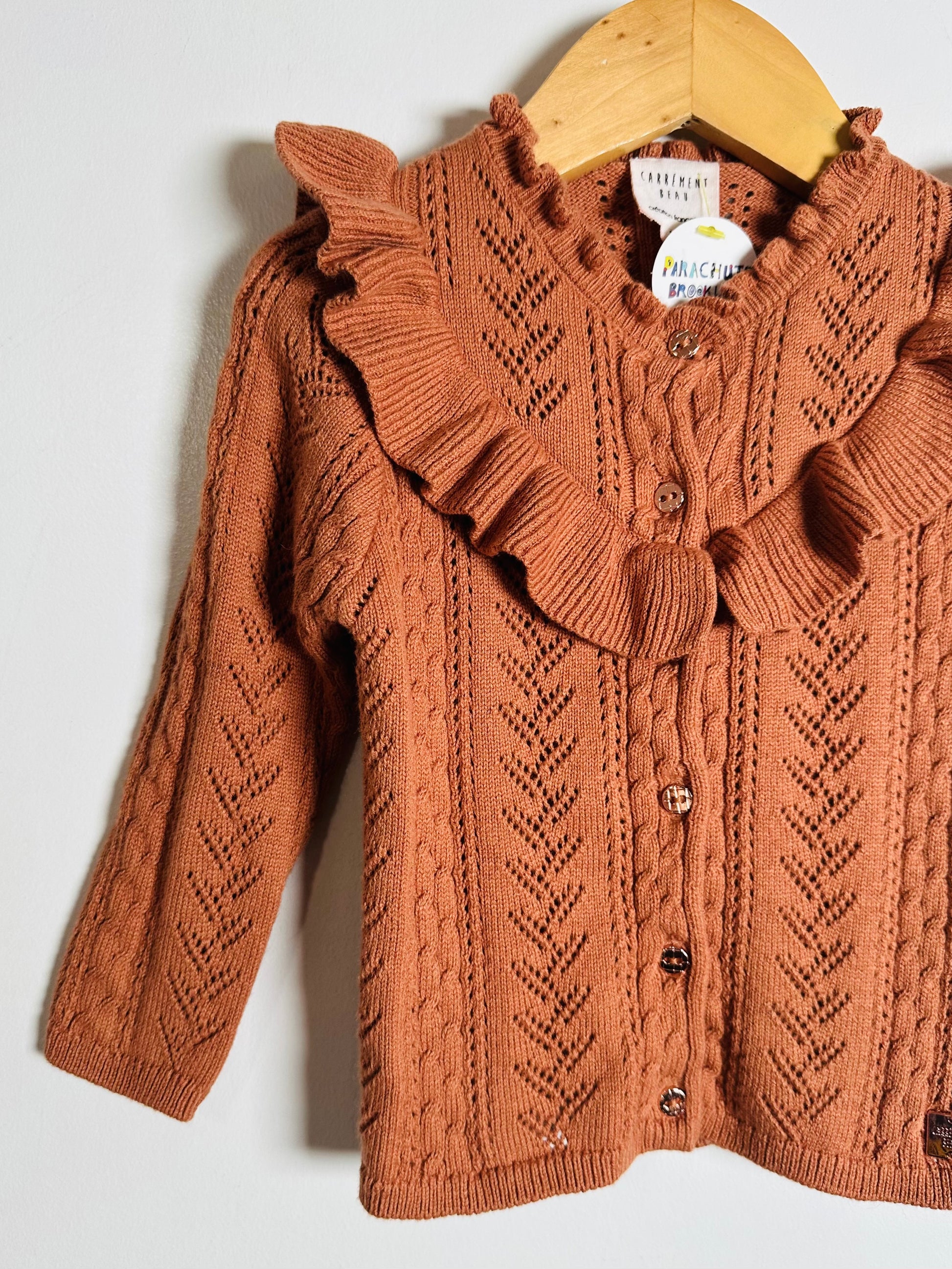 Brown knitted cardigan with ruffled collar on a wooden hanger against a white background