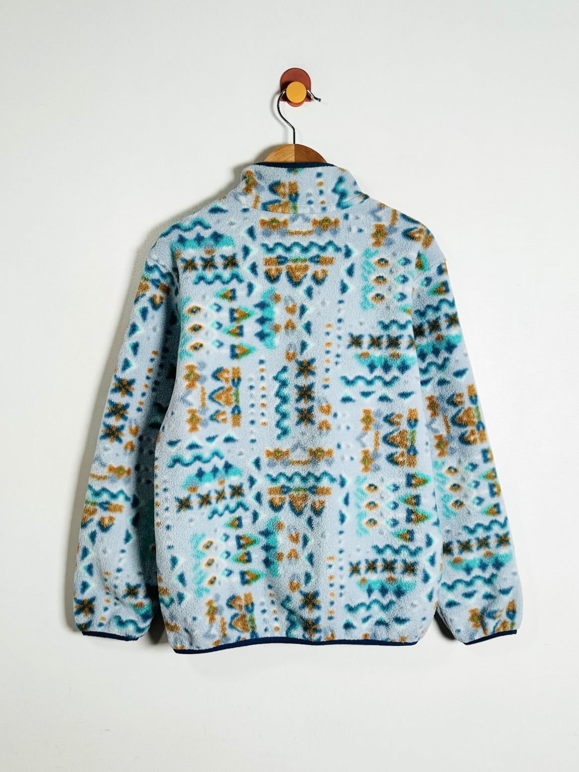 A light blue fleece jacket with a colorful geometric pattern in teal, orange, and yellow, hanging on a wooden hanger against a plain white wall.