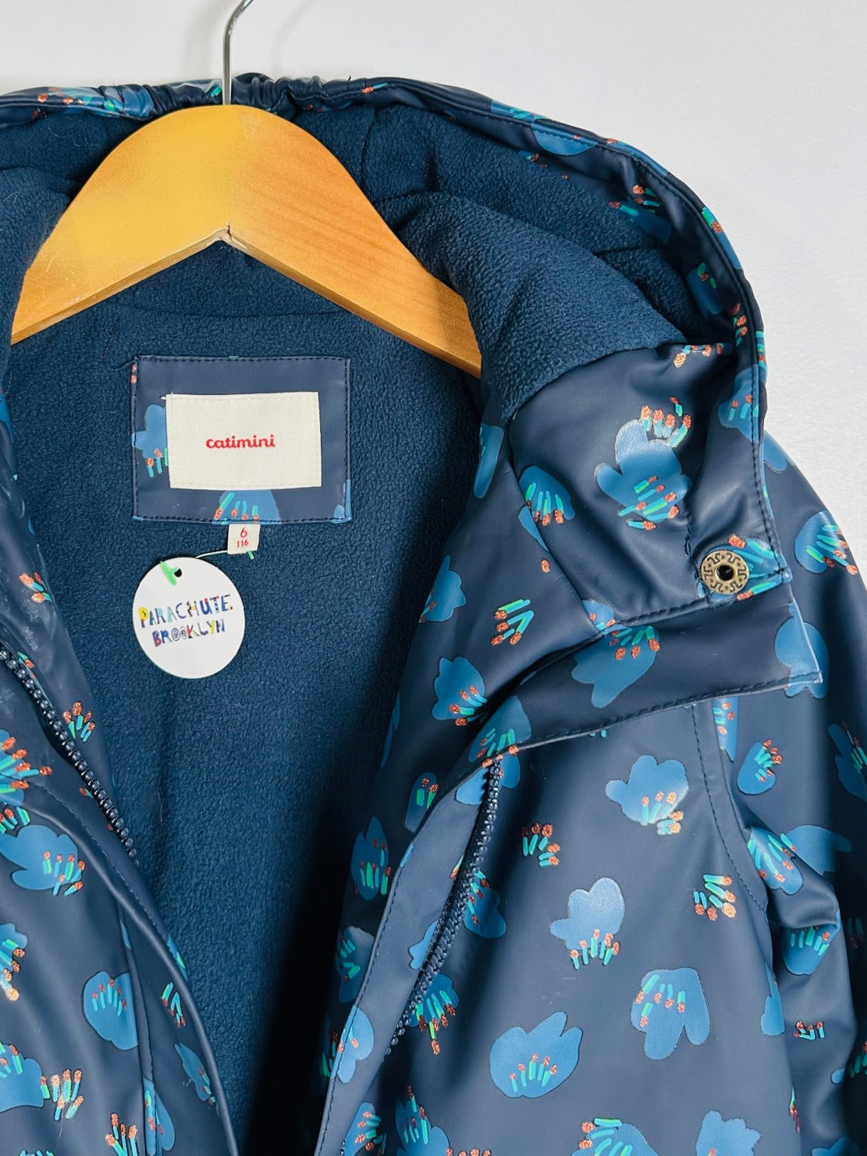 A close-up of a hooded jacket hanging on a wooden hanger against a plain light background. The jacket has a dark blue patterned outer shell with blue and teal cloud-like motifs, a dark blue fleece-lined hood and collar, and a visible interior label reading colmmini.