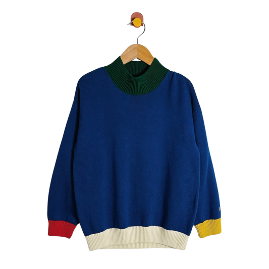 A blue knit sweater with a dark green mock neck, cream ribbed hem and cuffs, and red and yellow color-blocked accents at the cuffs, hanging on a wooden hanger against a white background.
