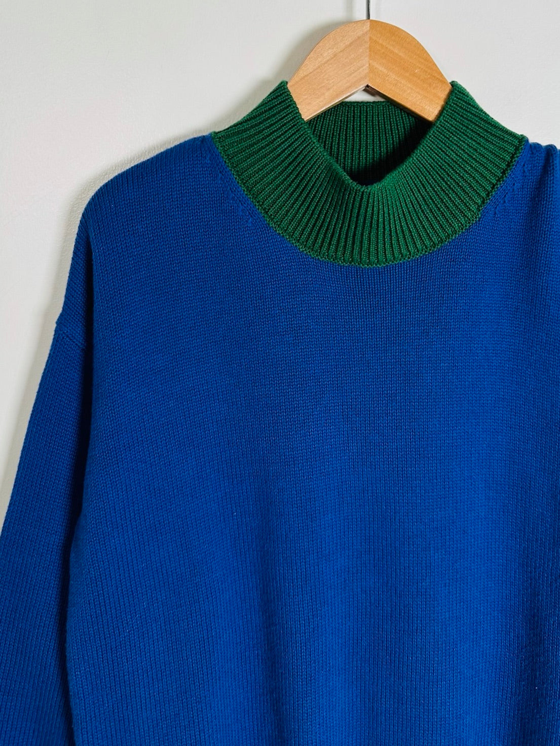 A blue knit sweater with a contrasting dark green mock neck collar, hanging on a wooden hanger against a plain light gray wall.