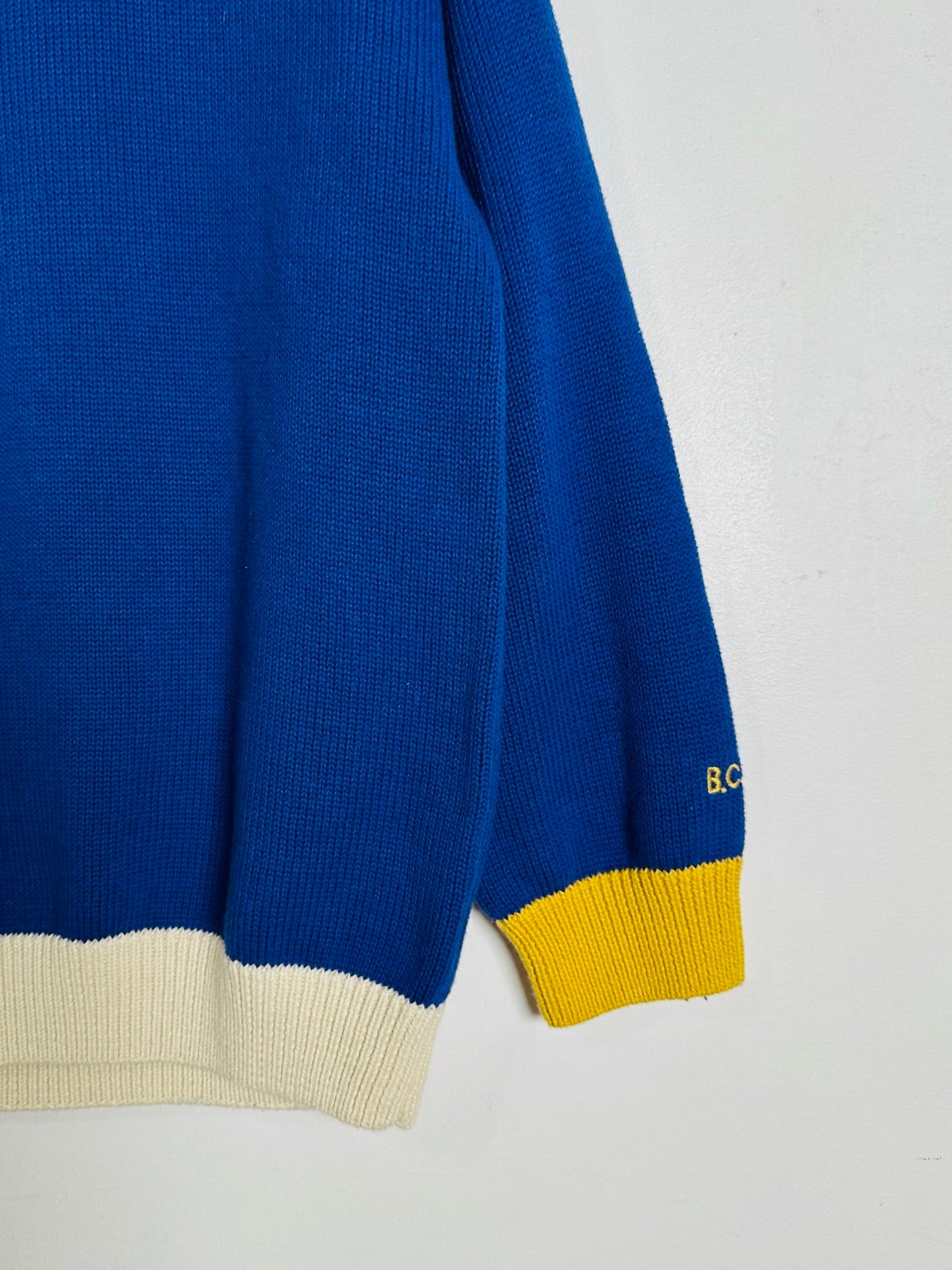 A close-up of a blue knit sweater sleeve with a cream ribbed hem and a yellow ribbed cuff, featuring small yellow embroidered letters near the cuff.