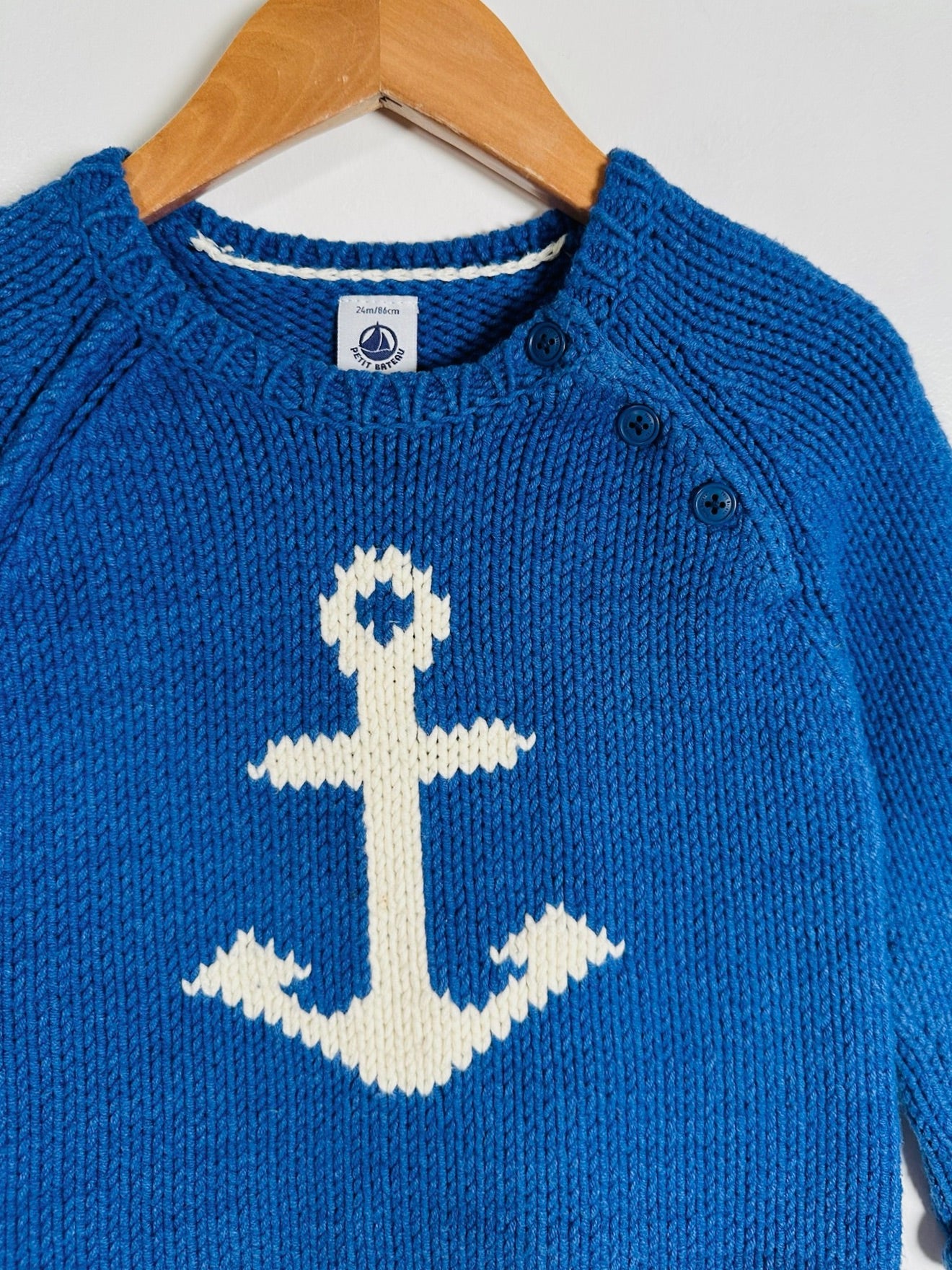 A close-up of a blue knit sweater hanging on a wooden hanger against a light gray background. The sweater features a large white knitted anchor motif on the front and three dark buttons near the collar.