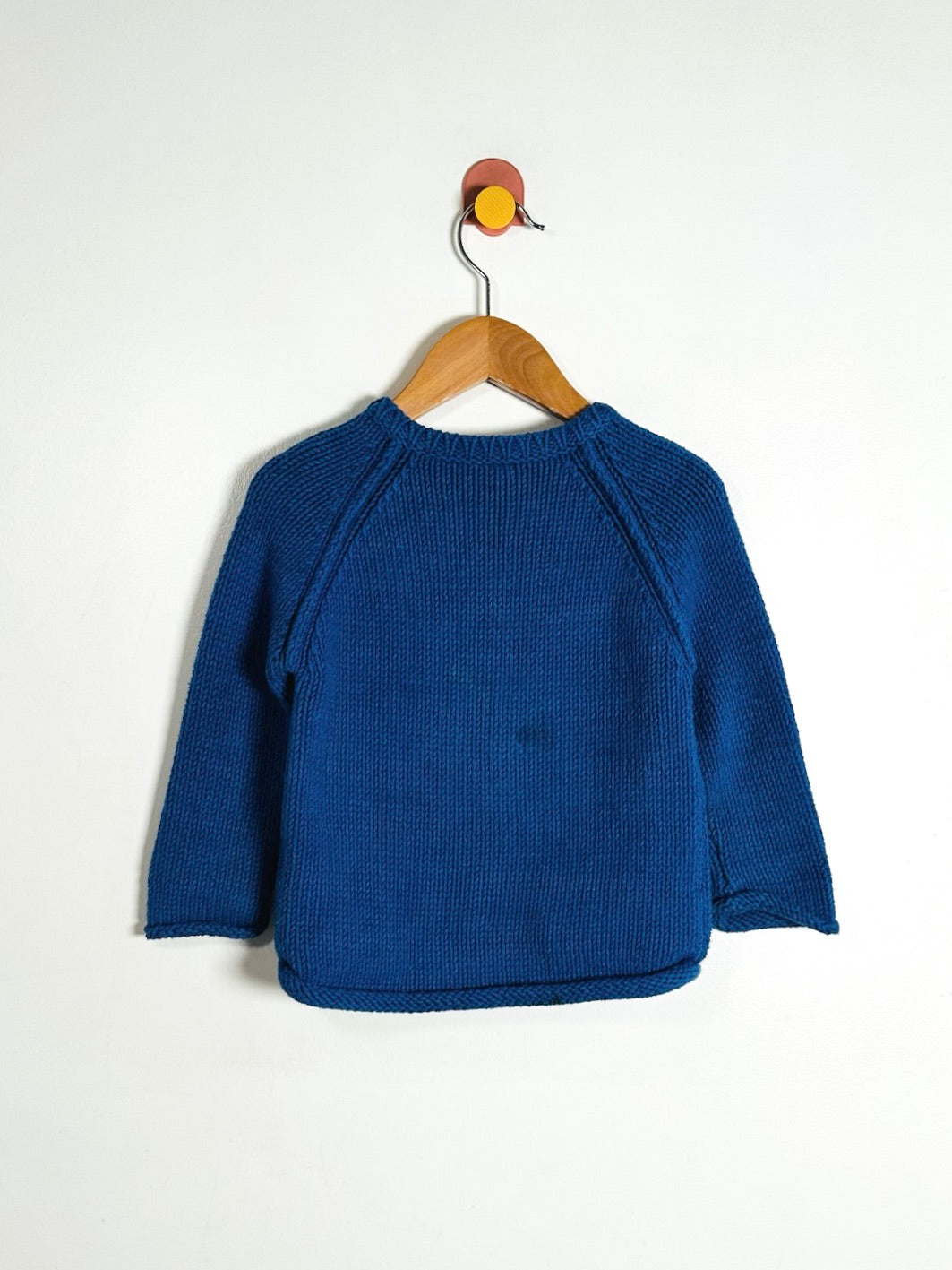 A royal blue knit raglan sweater hanging on a wooden hanger against a plain white wall.