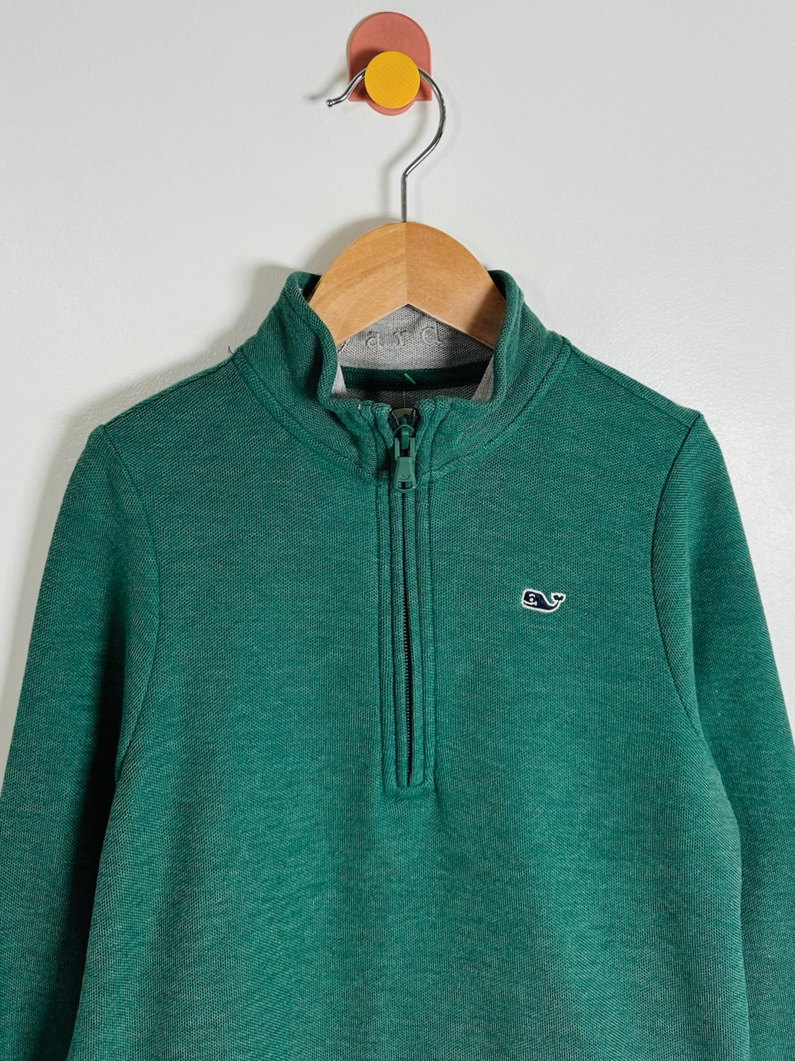 A green heathered quarter-zip pullover sweatshirt hanging on a wooden hanger against a plain light gray wall. The sweatshirt features a small white embroidered whale logo on the left chest.