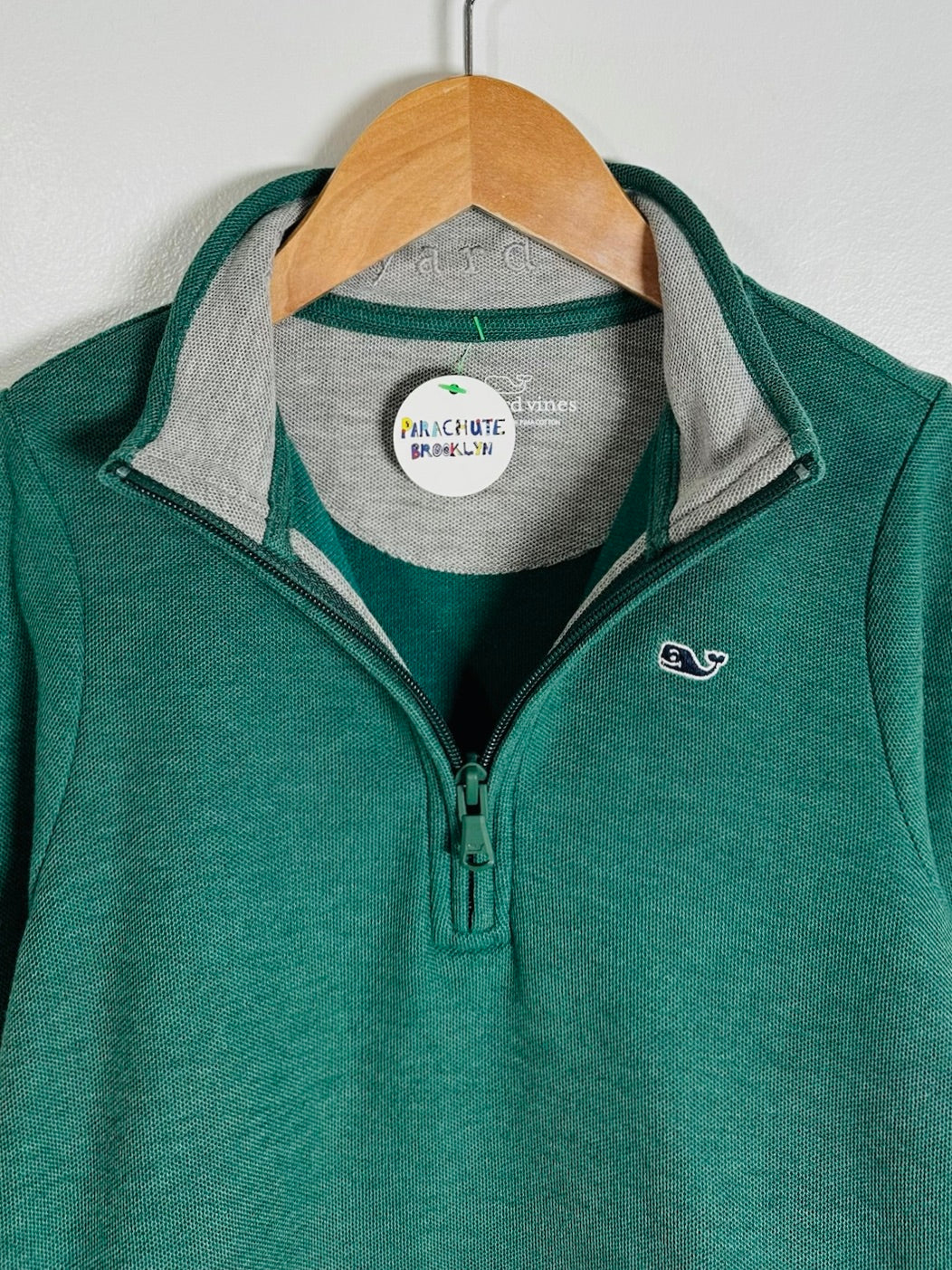 A green quarter-zip pullover sweater with a gray inner collar, hanging on a wooden hanger against a light gray wall. The brand label reads 'Natchezta Apparel' and the chest features a small white whale logo.