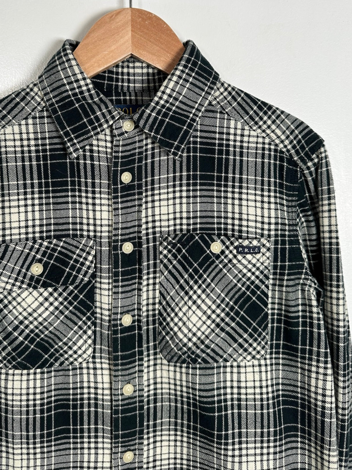 A black and off-white plaid button-down shirt hanging on a wooden hanger against a plain light gray wall. The shirt features a classic collar, front button closure, and two chest pockets.