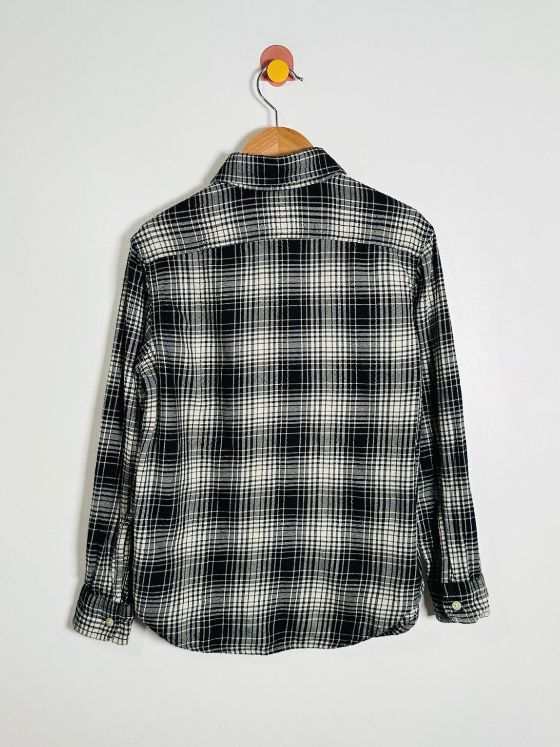 A black and white plaid long-sleeve button-up shirt hanging on a wooden hanger against a plain light gray wall.