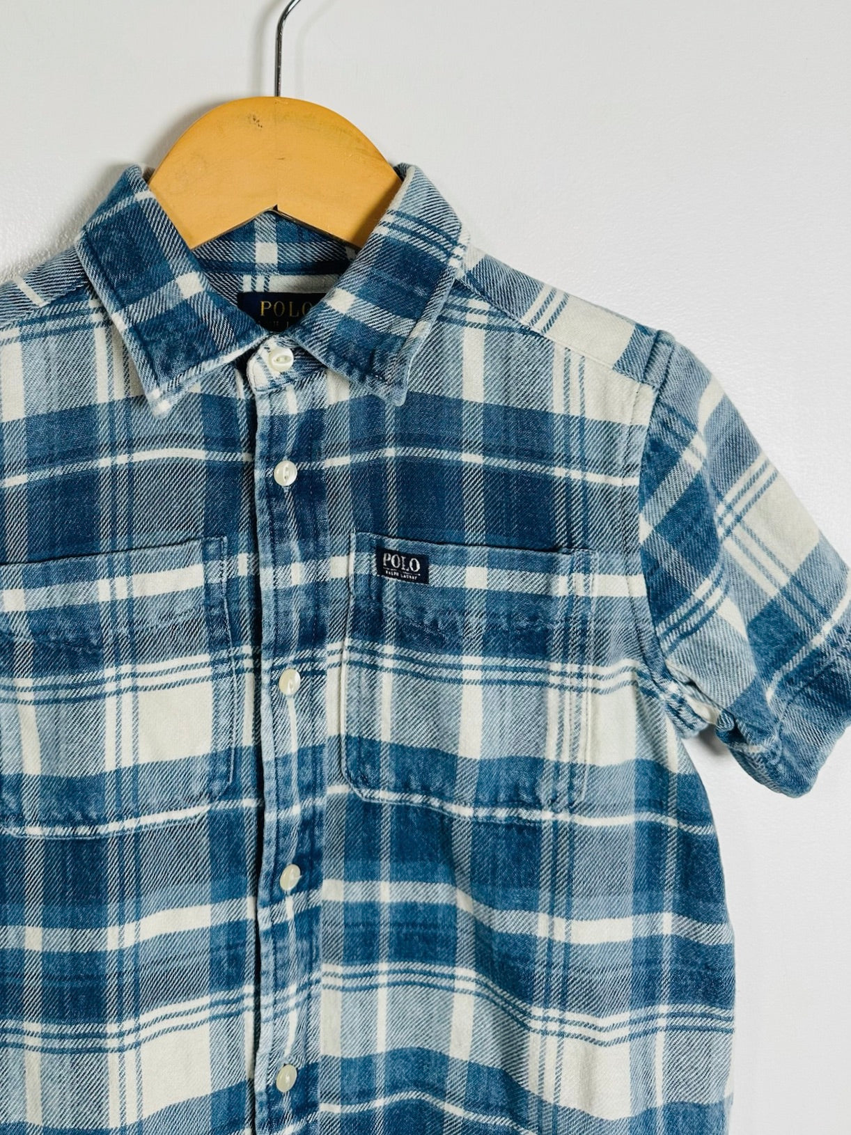 A short-sleeve button-up plaid shirt in blue and white, hanging on a wooden hanger against a plain light gray wall. A small chest label reads Polo.