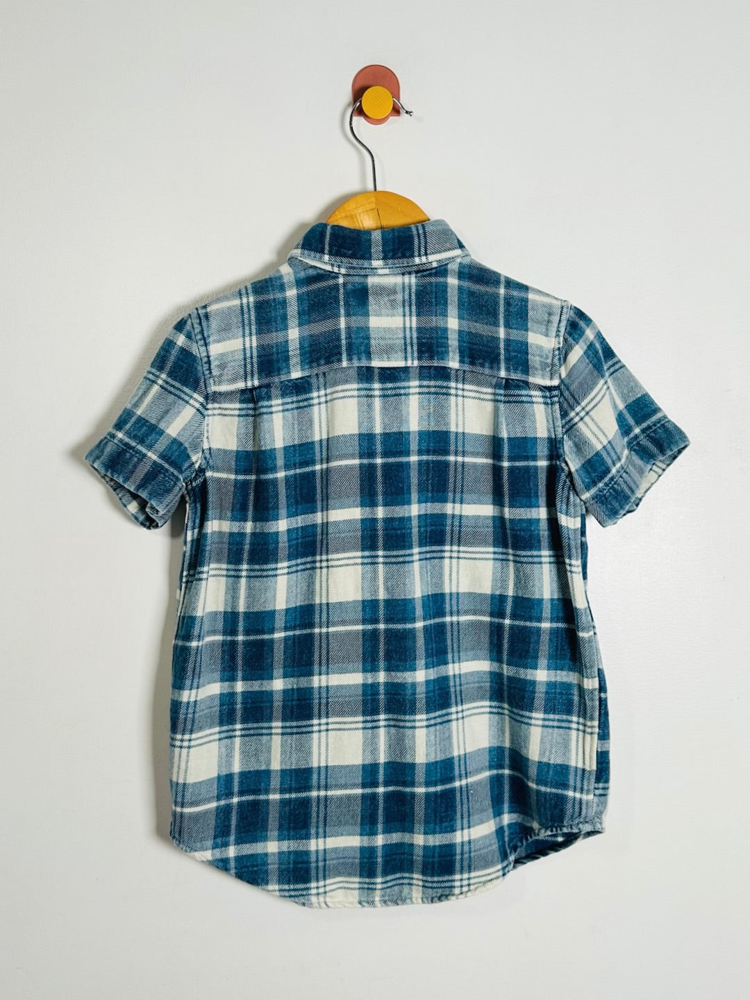 A short-sleeve plaid button-up shirt in blue and white, hanging on a wooden hanger with a red and yellow wall hook against a plain light gray wall.
