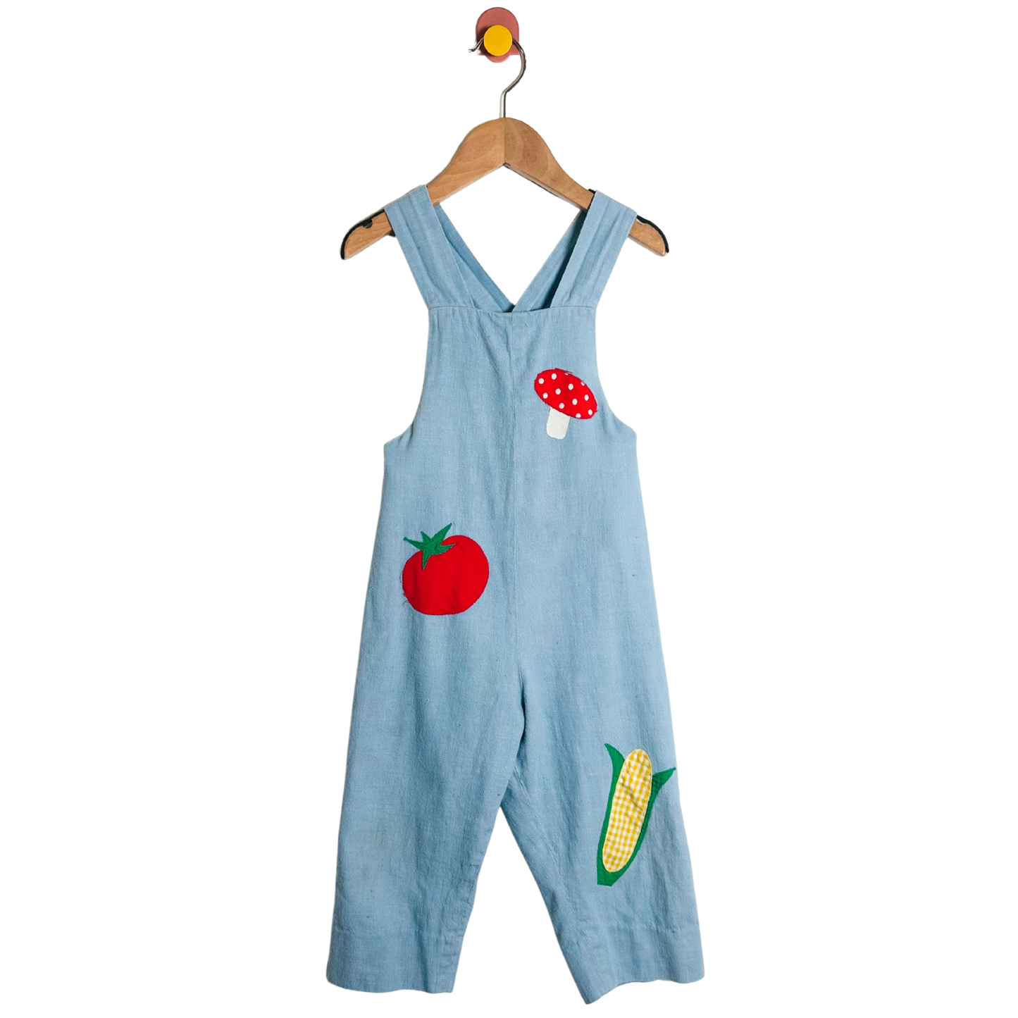 Light blue overalls with tomato and corn appliqués on a hanger against a white background