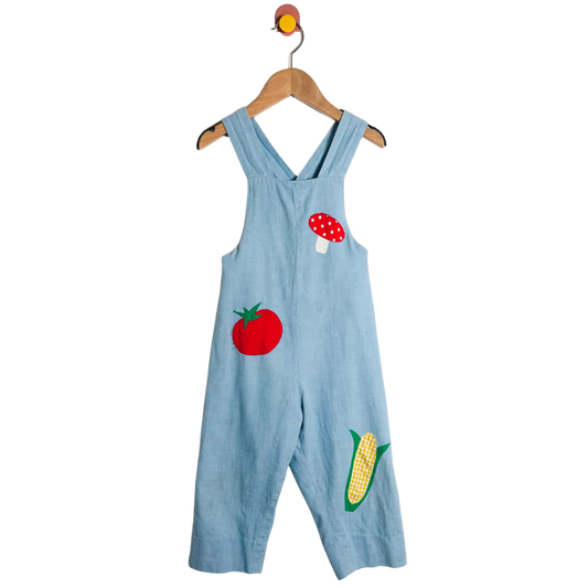 Light blue overalls with tomato and corn appliqués on a hanger against a white background