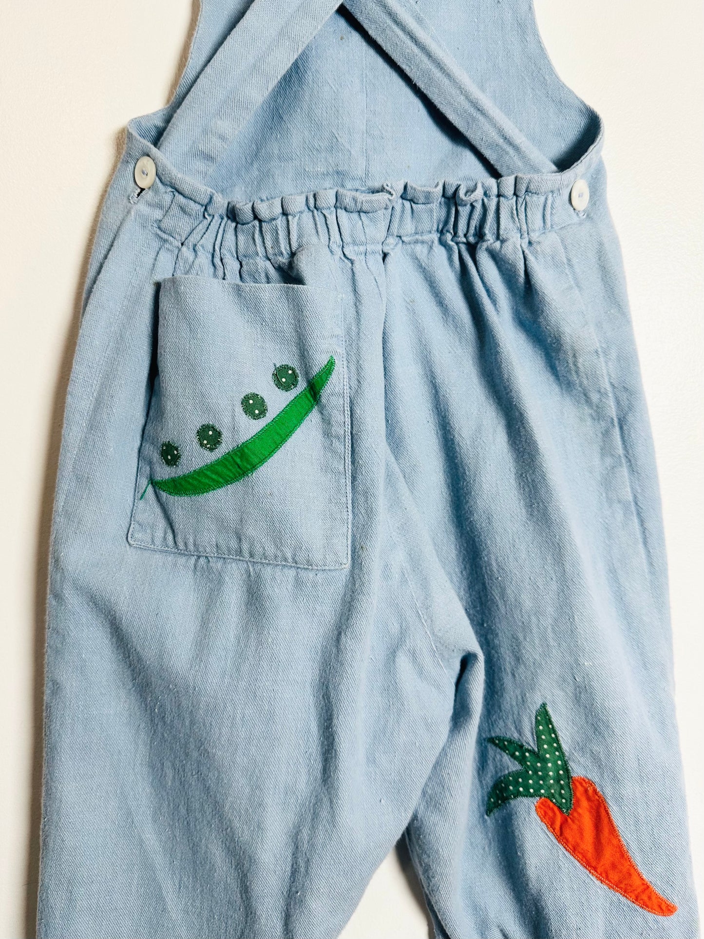 Light blue denim overalls with embroidered green bean and orange carrot designs on a beige background.