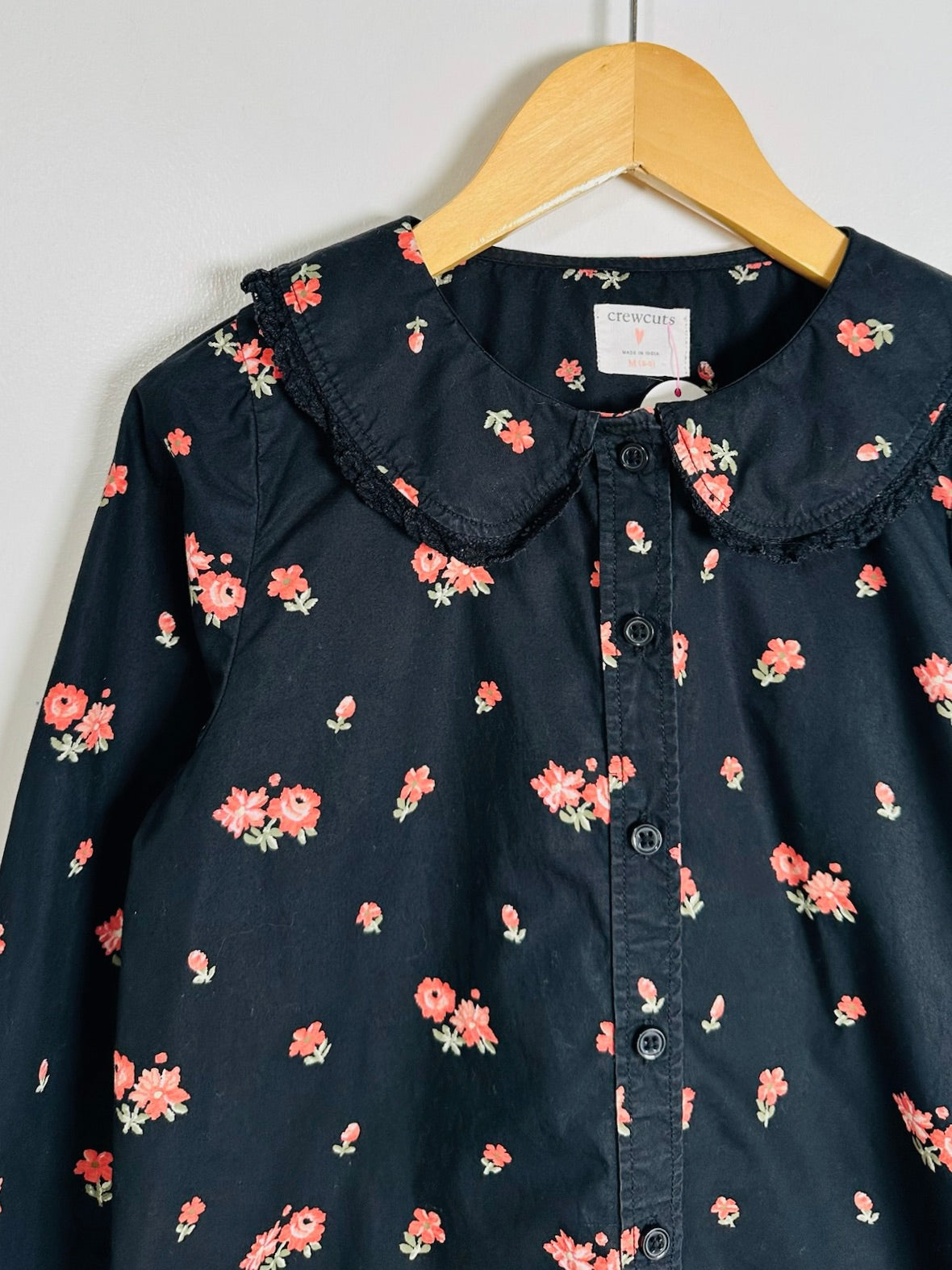 A dark navy blue long-sleeve blouse with a pink and green floral print, featuring a ruffled Peter Pan collar and a button-down front, hanging on a wooden hanger against a plain light gray wall.